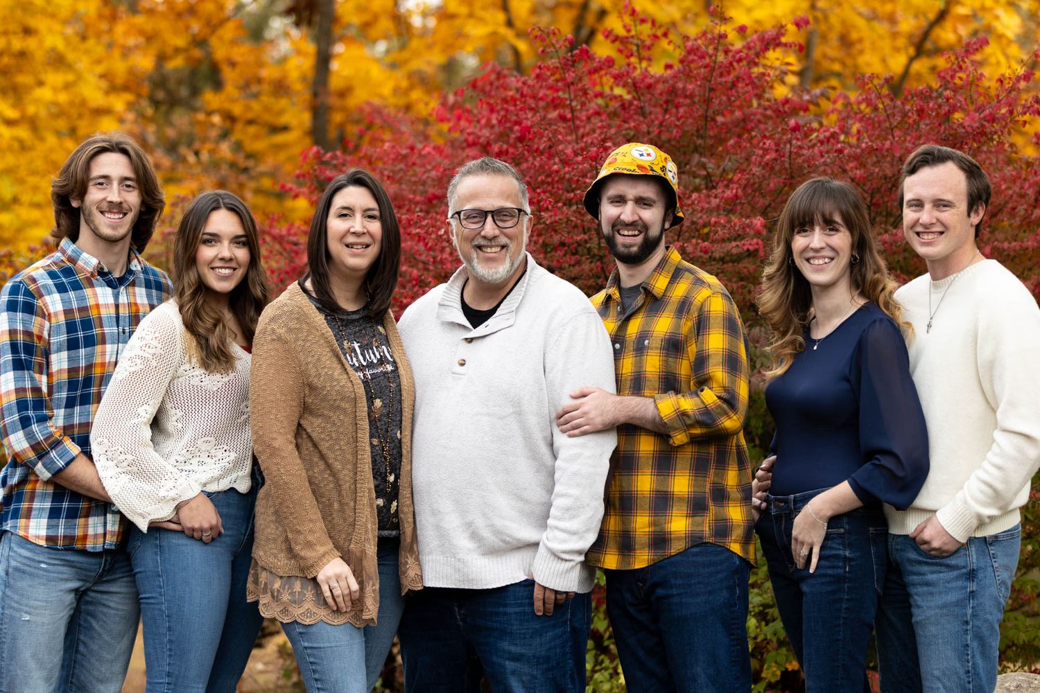 Happy family of seven adults posing together outdoors surrounded by vibrant fall foliage colors.