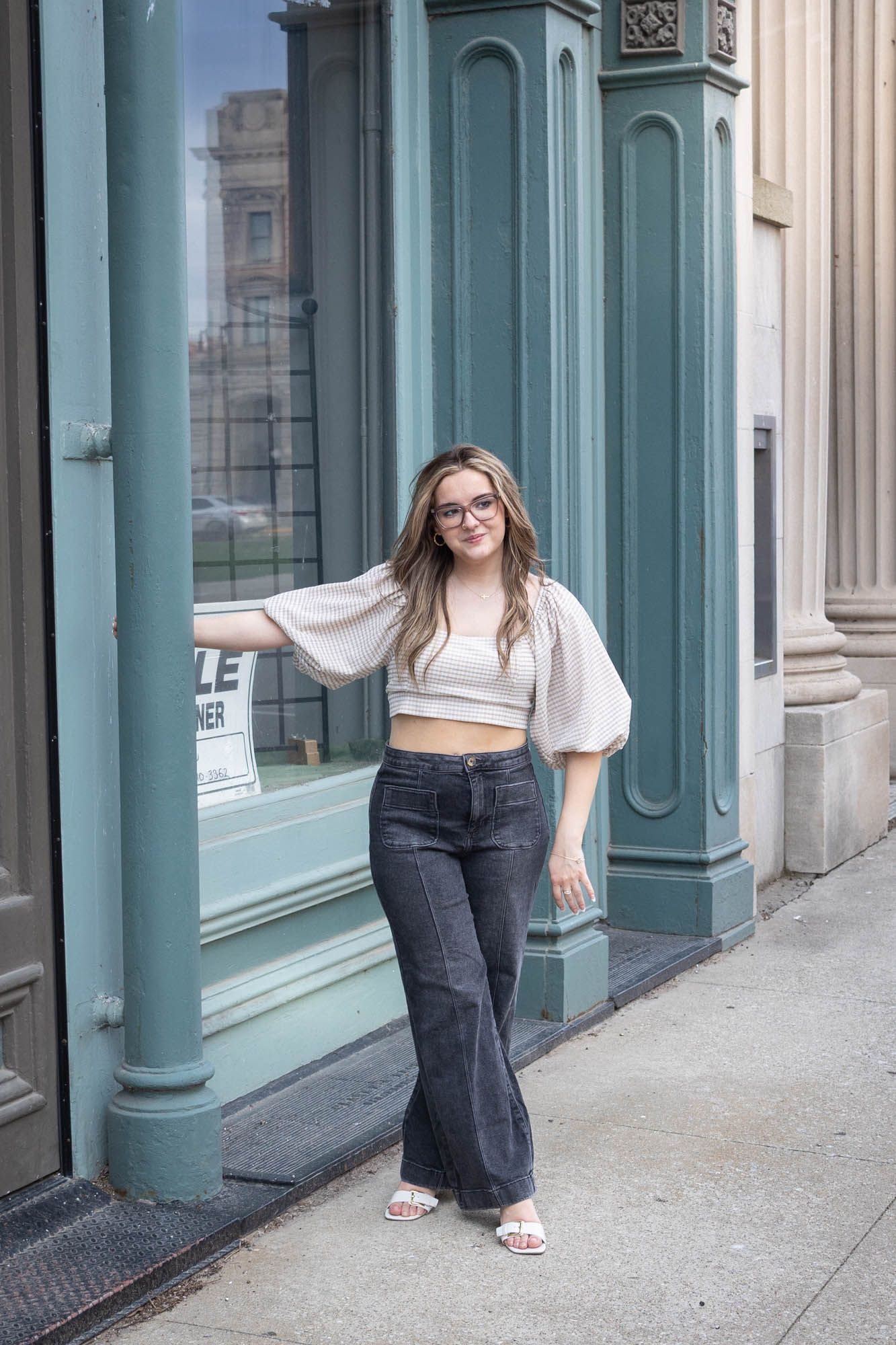 Confident young woman in white crop top and dark flare jeans posing by green storefront columns.