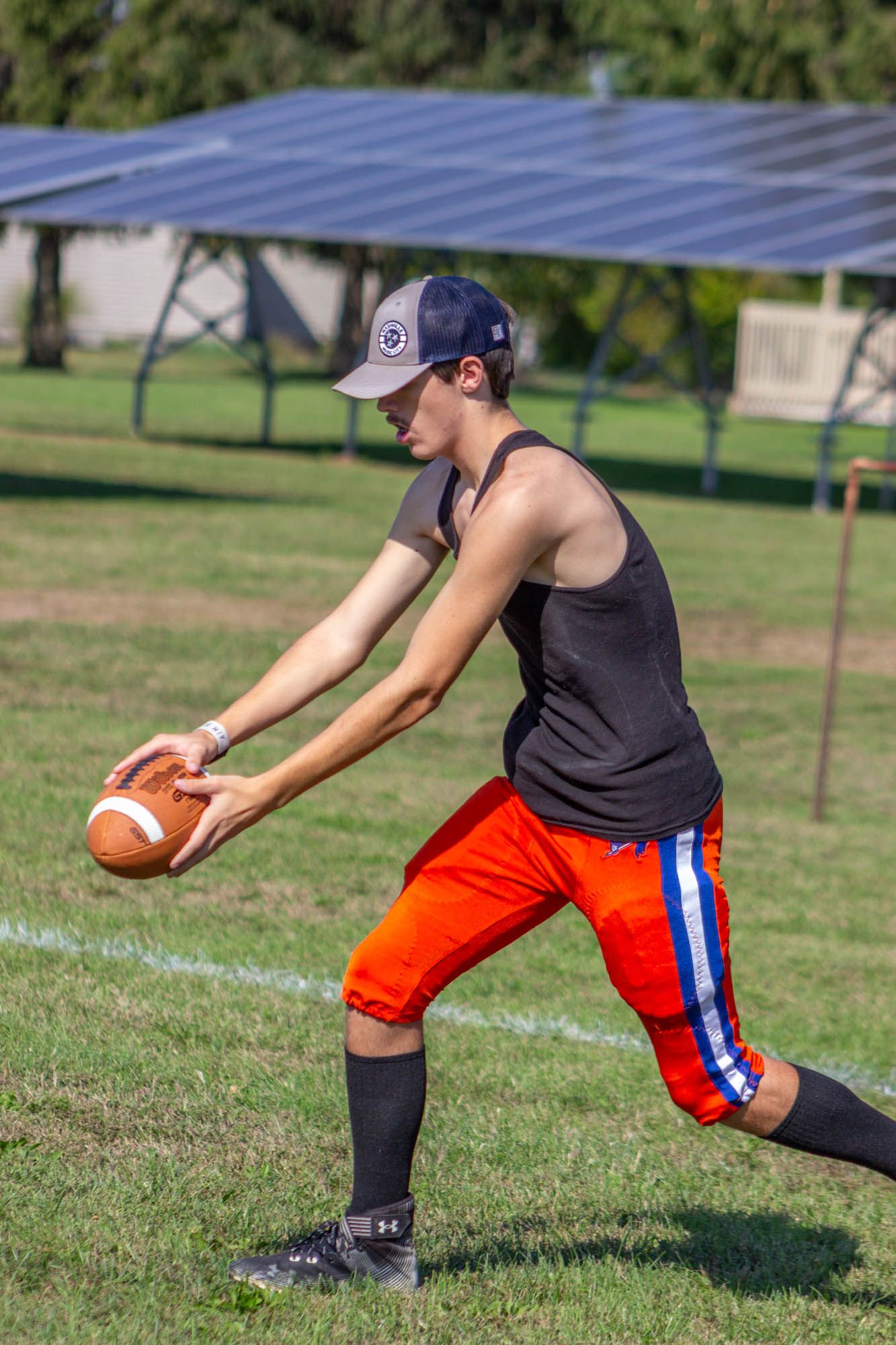 Young football player in black tank top and orange shorts practices snapping the ball on a sunny field.