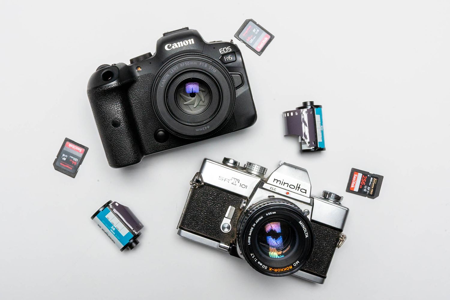 Flat lay of Canon EOS DSLR and Minolta film camera with memory cards and film rolls on white surface.
