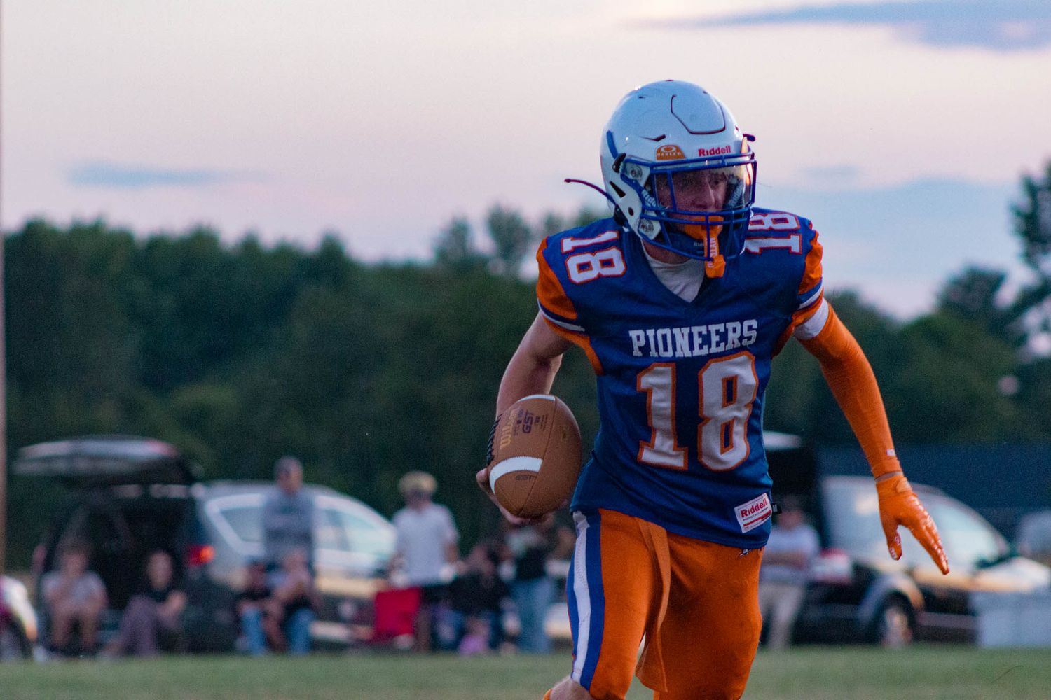 Pioneer football player number 18 in blue and orange uniform carries the ball during an evening game.