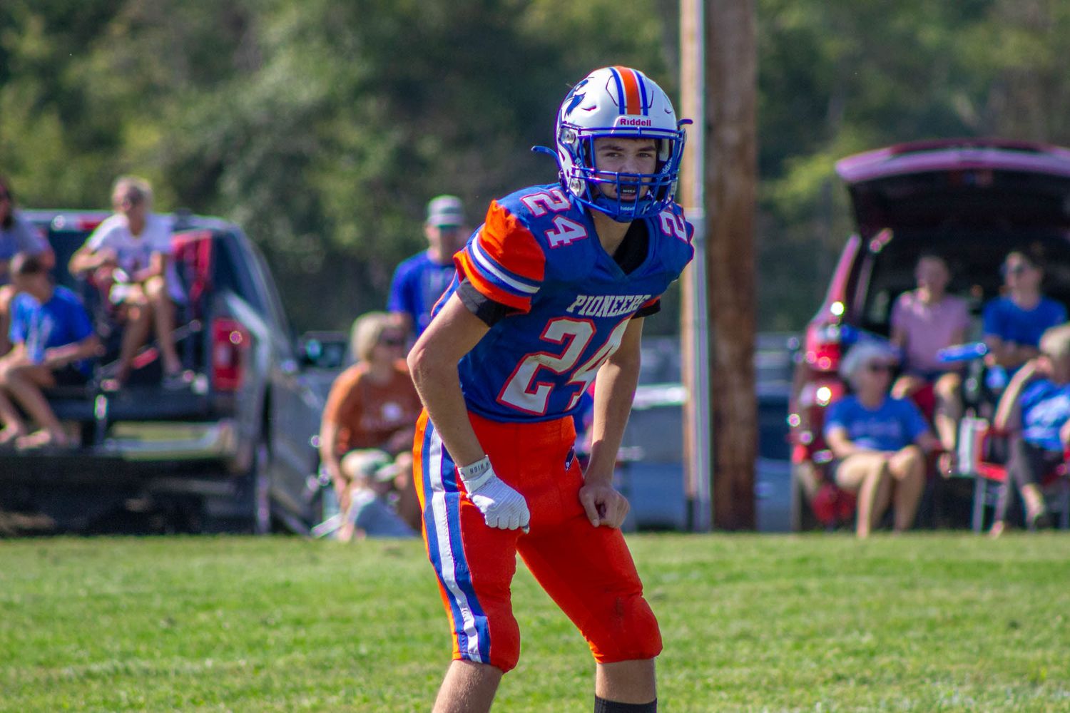 A high school football player in blue and orange uniform number 24 stands ready on the field during a sunny game.