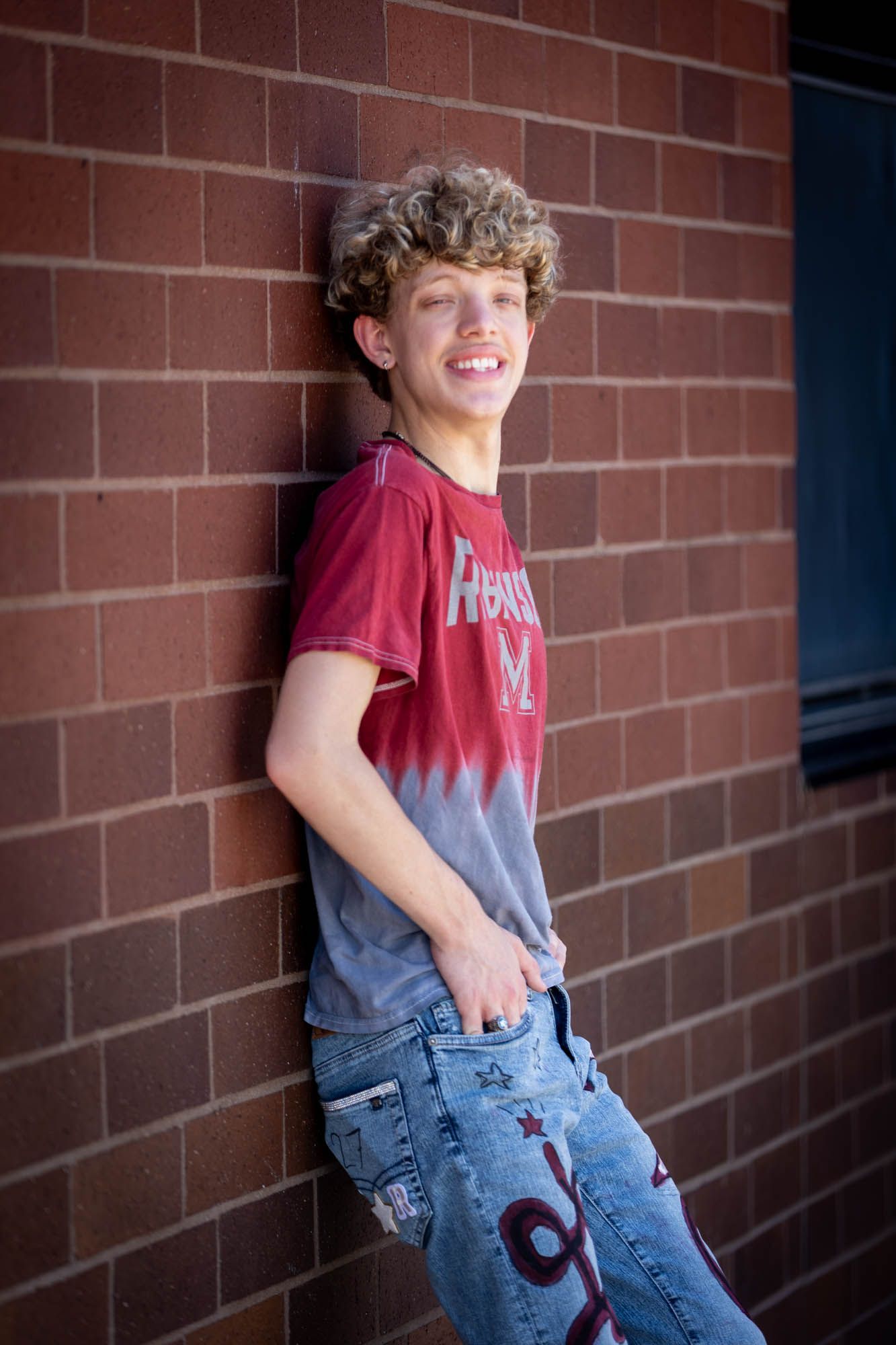 Smiling teenage boy with curly hair leaning against a brick wall wearing a red graphic tee and distressed jeans.