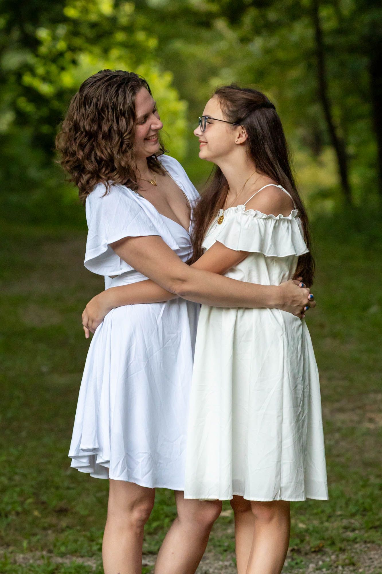 Two women in white dresses embracing and smiling at each other outdoors in a lush green park setting.