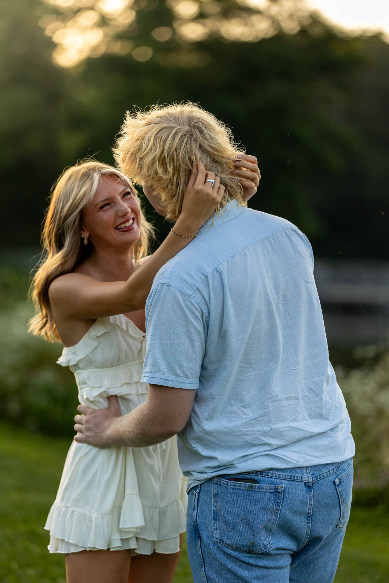 A smiling blonde woman in a white dress playfully touches a man's hair outdoors at golden hour.
