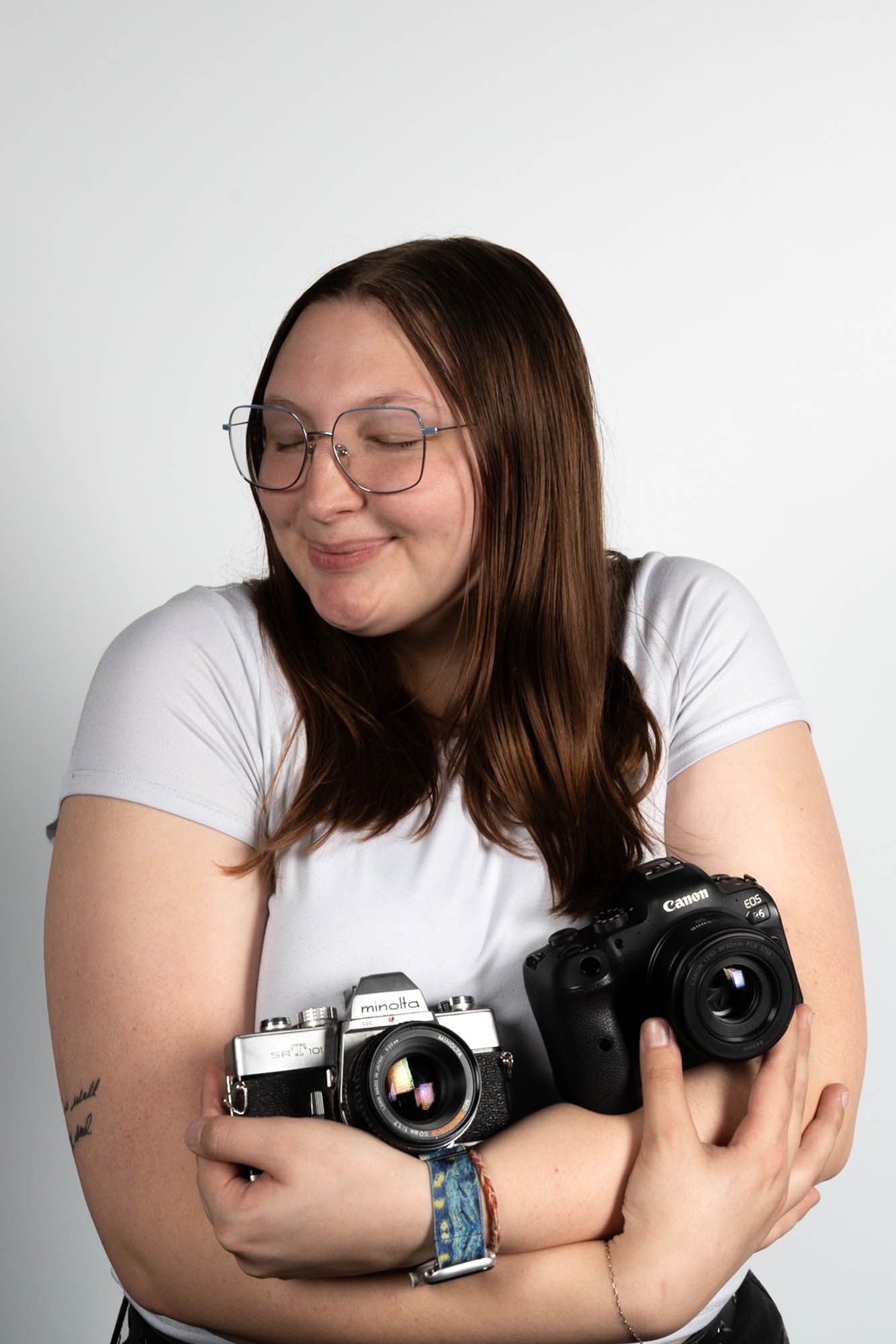 Young woman in white t-shirt smiling while cradling two cameras, a Canon DSLR and film camera.