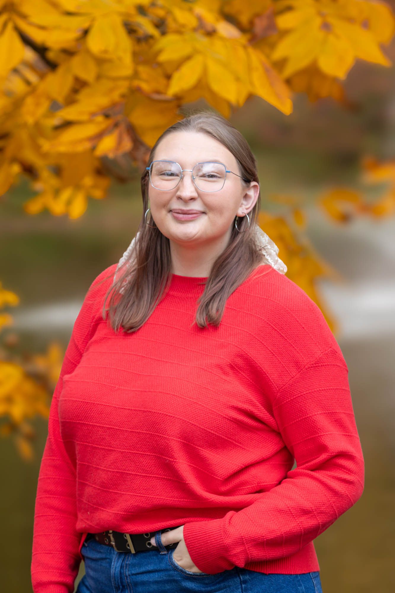 Young woman in red sweater and glasses smiling outdoors with golden autumn leaves and pond in background.