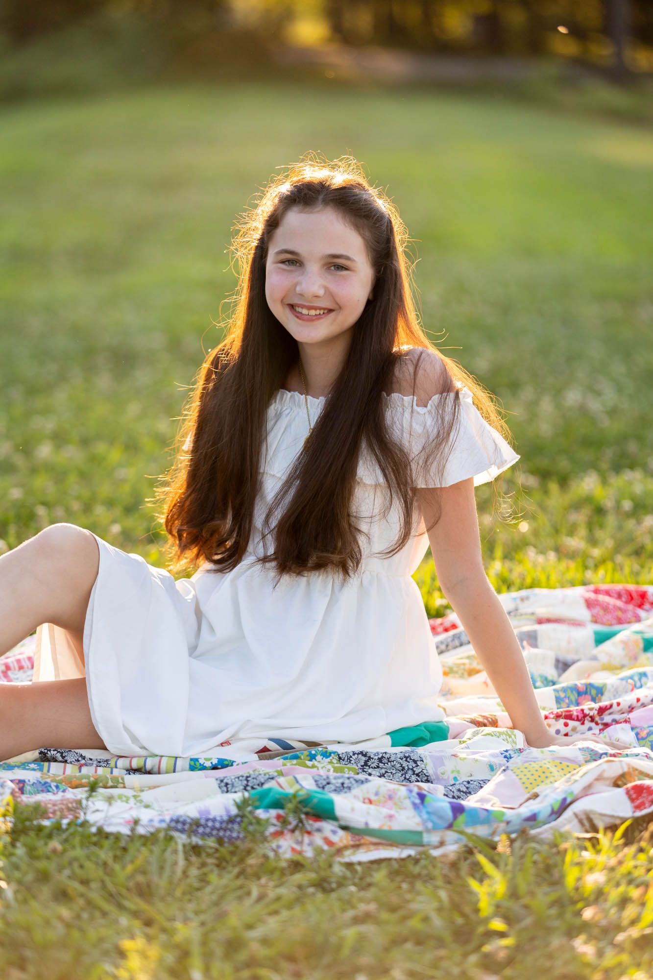 Smiling teenage girl with long brown hair sitting on a colorful quilt blanket in a sunny green park wearing a white dress.