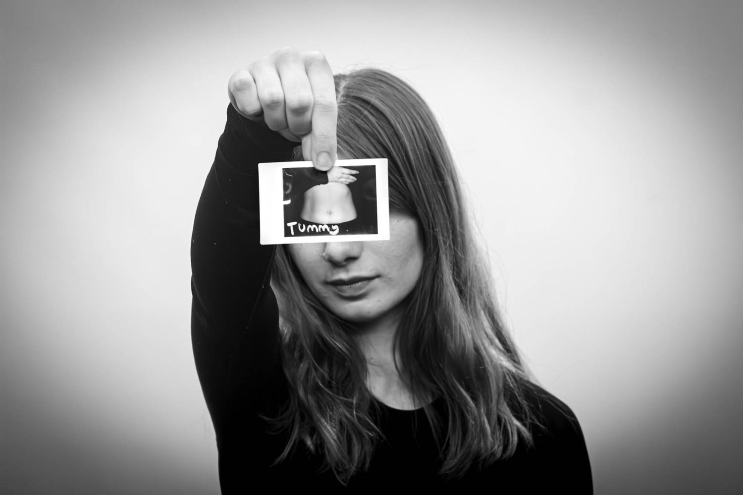 Young woman holding a labeled Polaroid photo covering her face in a black and white portrait.