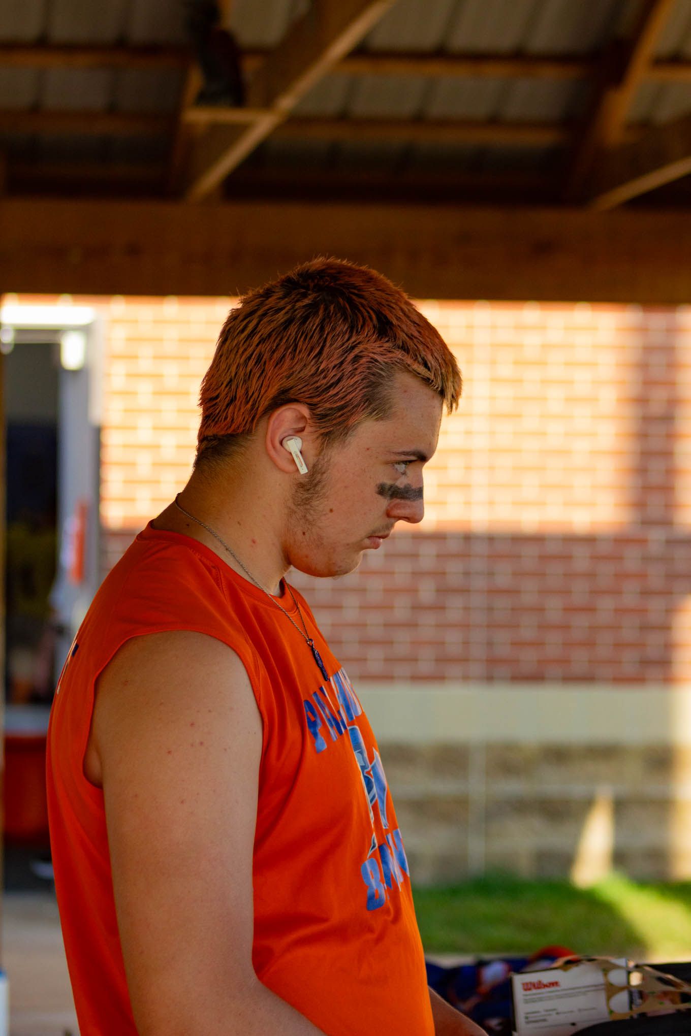 Young man with orange-dyed hair wearing an orange sleeveless shirt with earbuds outdoors.