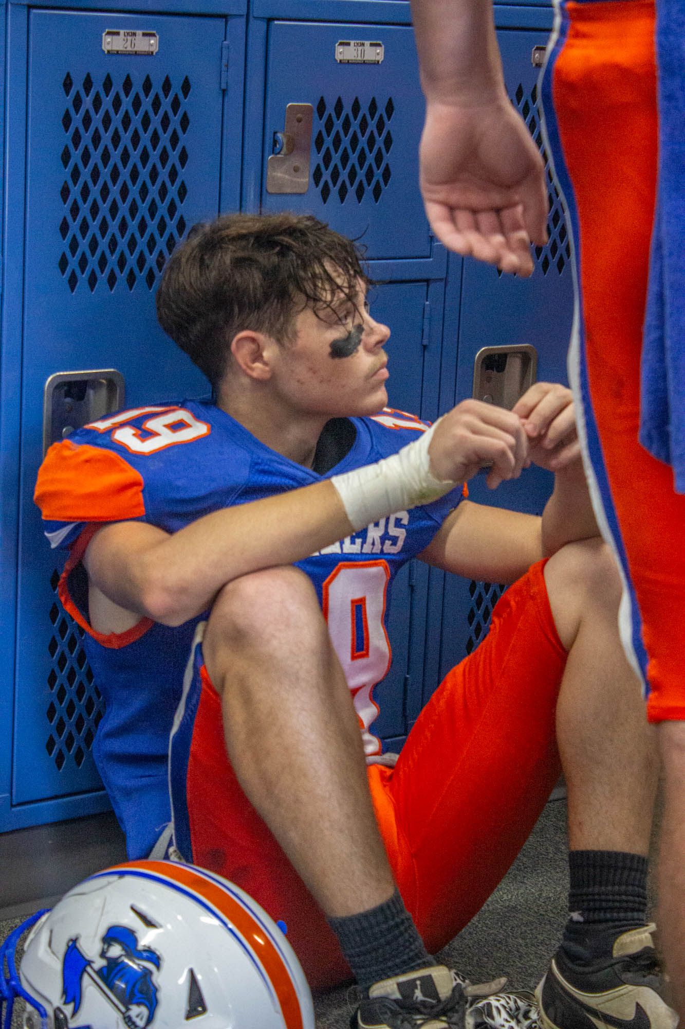 Tired Pioneers football player sits against blue lockers in locker room after a game.