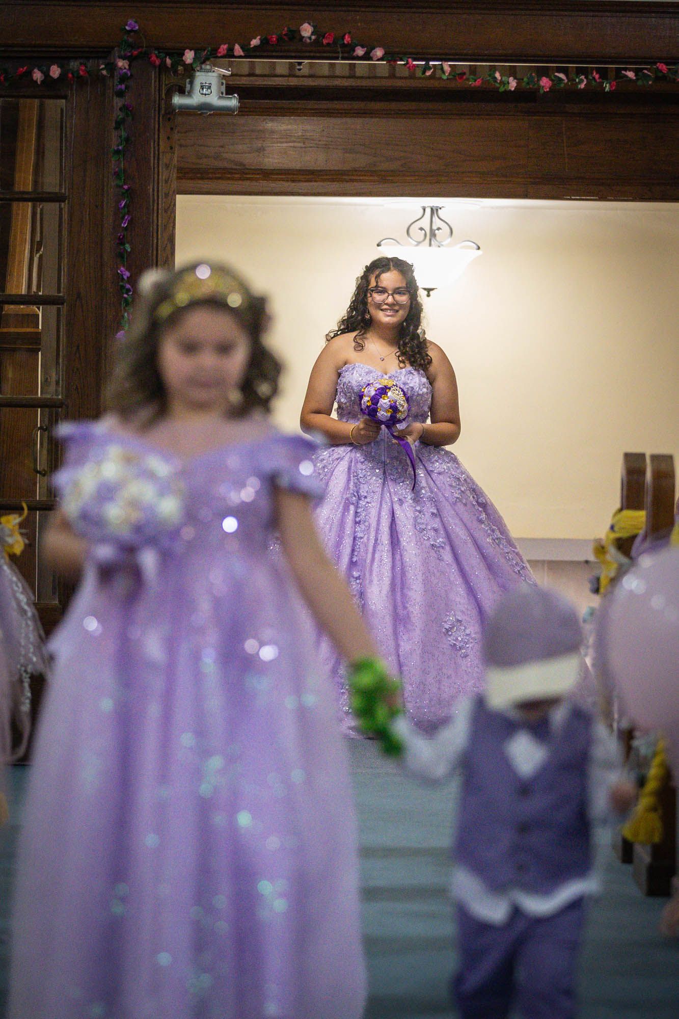 Young woman in a sparkling lavender quinceañera ball gown walks down the aisle with flower girl and ring bearer.