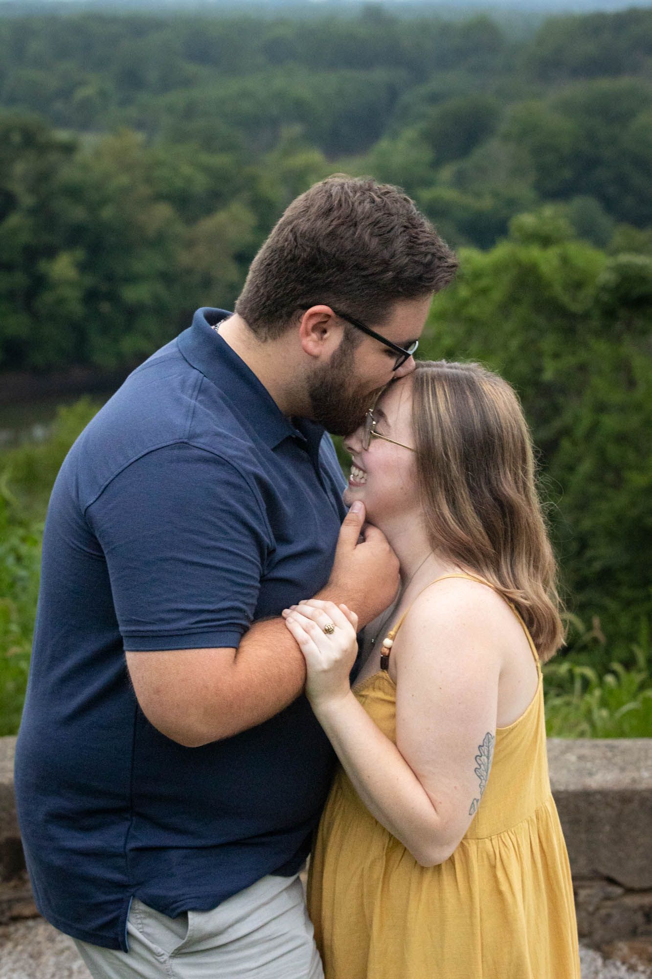 Couple sharing a romantic moment outdoors, man kissing woman's forehead while holding hands with lush green scenery behind them.