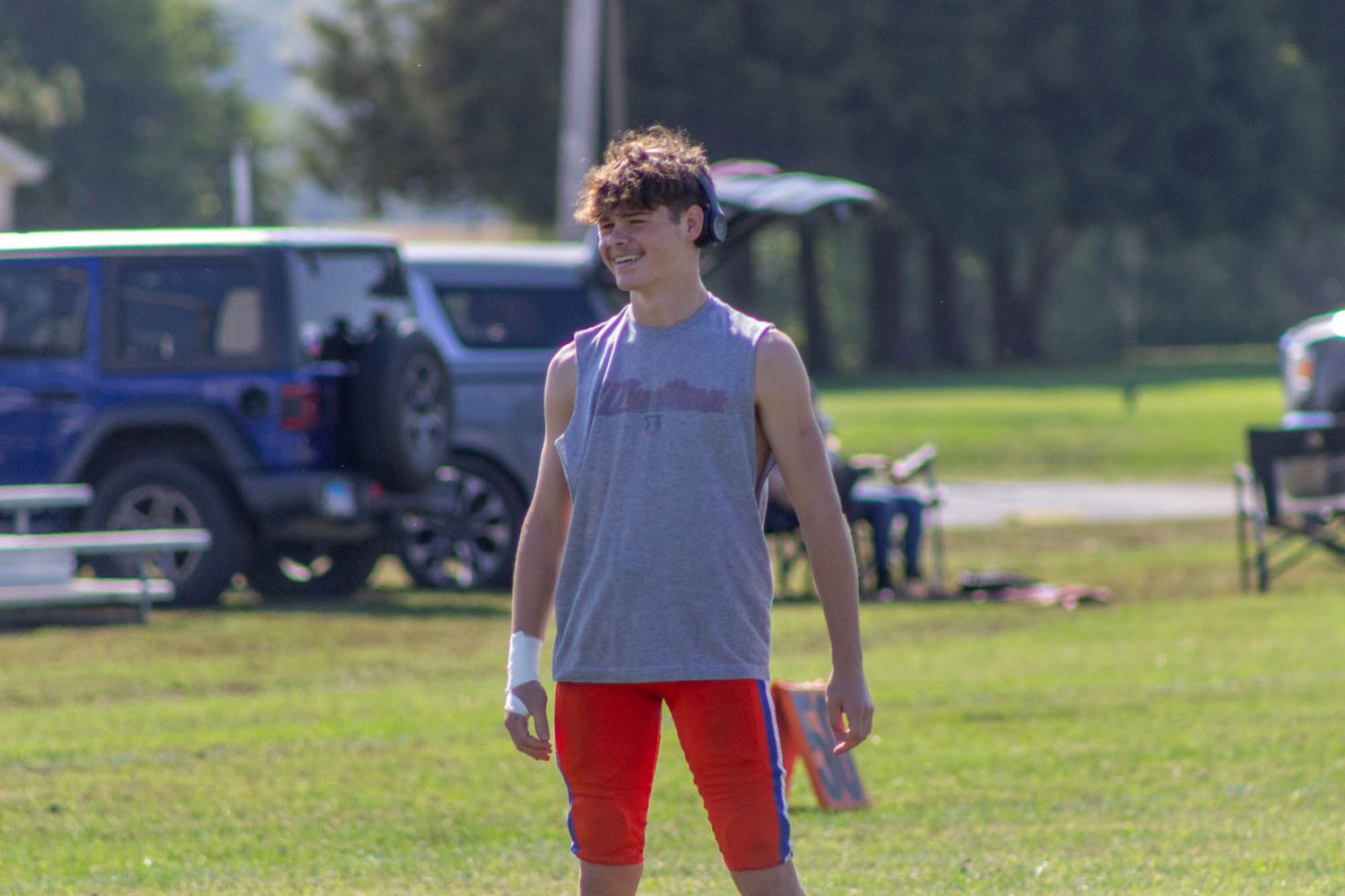 Young athlete in gray sleeveless shirt and red shorts stands on a sunny green practice field.