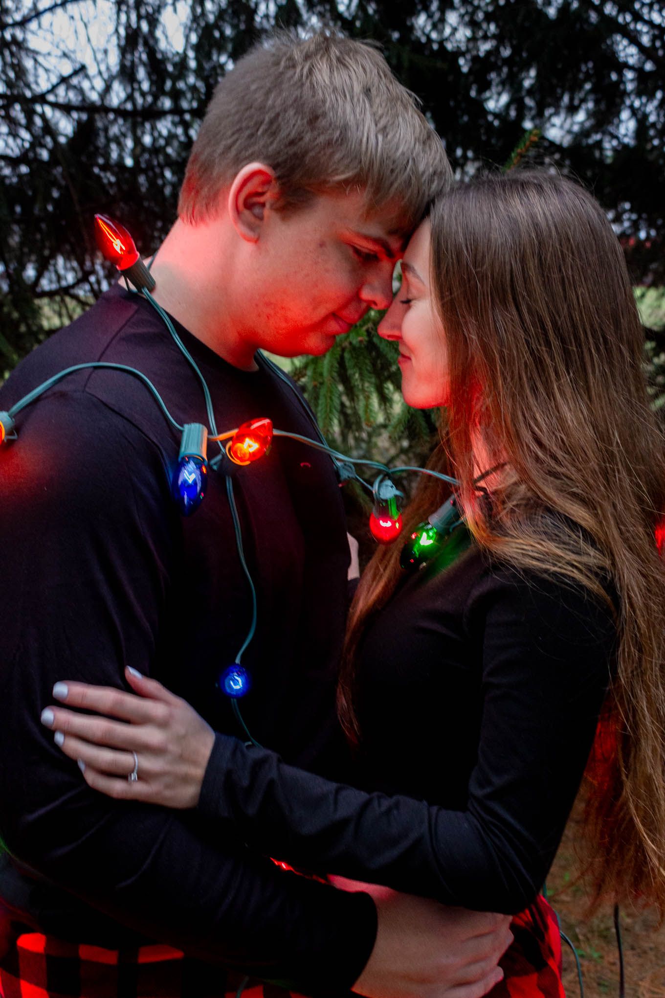 Young couple embracing and touching foreheads wrapped in glowing colorful Christmas lights outdoors.
