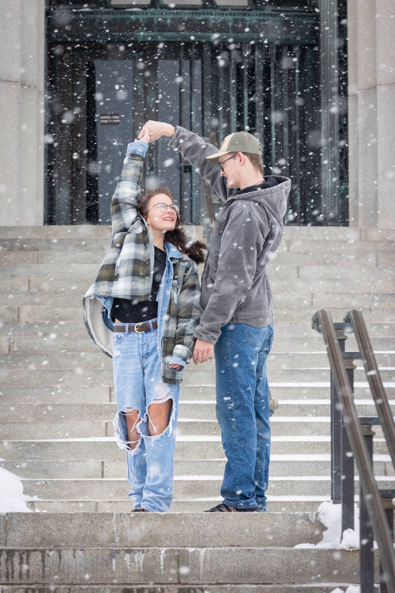 Young couple dancing playfully on snowy steps during a winter snowfall outdoors.