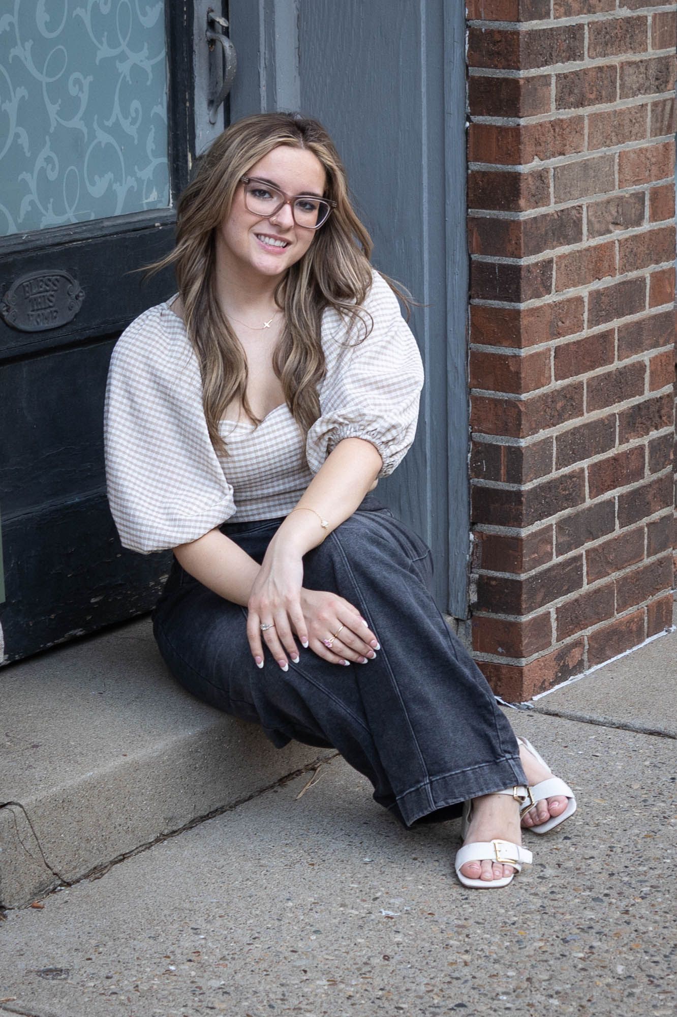 Smiling young woman with glasses in crop top and wide-leg jeans sitting on brick building steps.