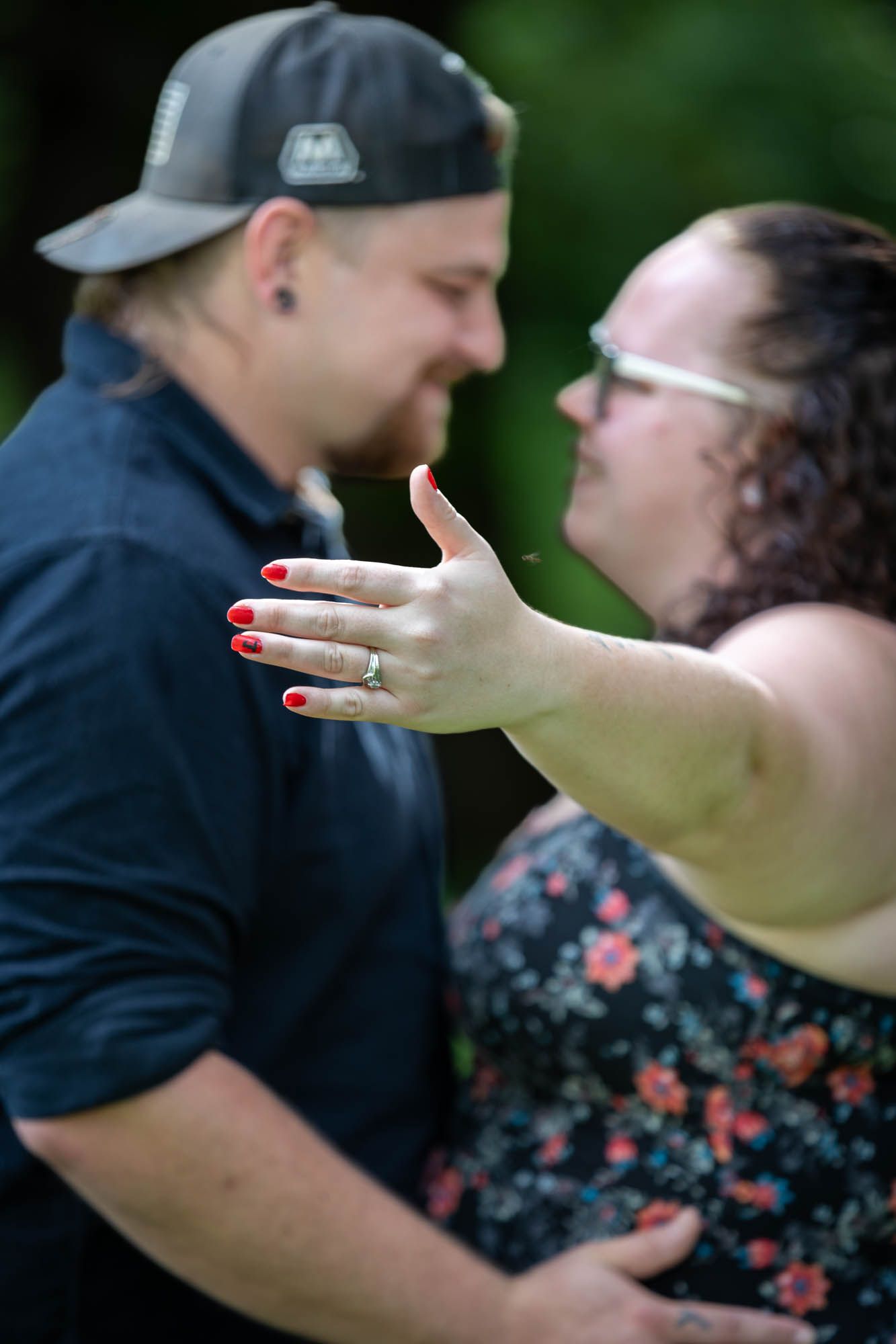 Couple sharing a romantic moment, woman displaying engagement ring with red nails toward camera outdoors.