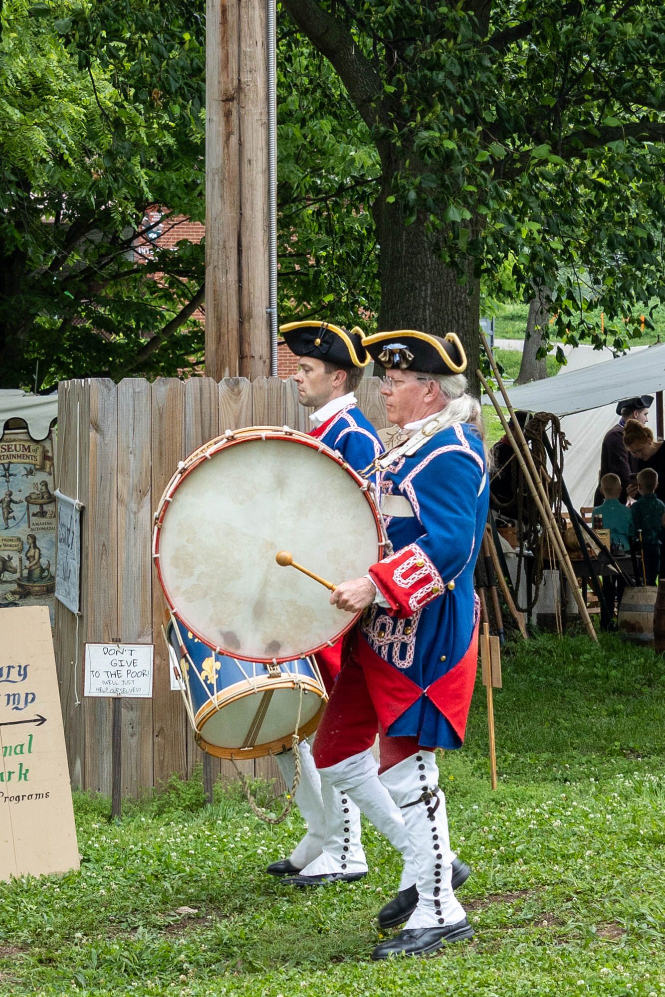 Colonial reenactors in blue and red uniforms march while playing a large bass drum at a historical festival.