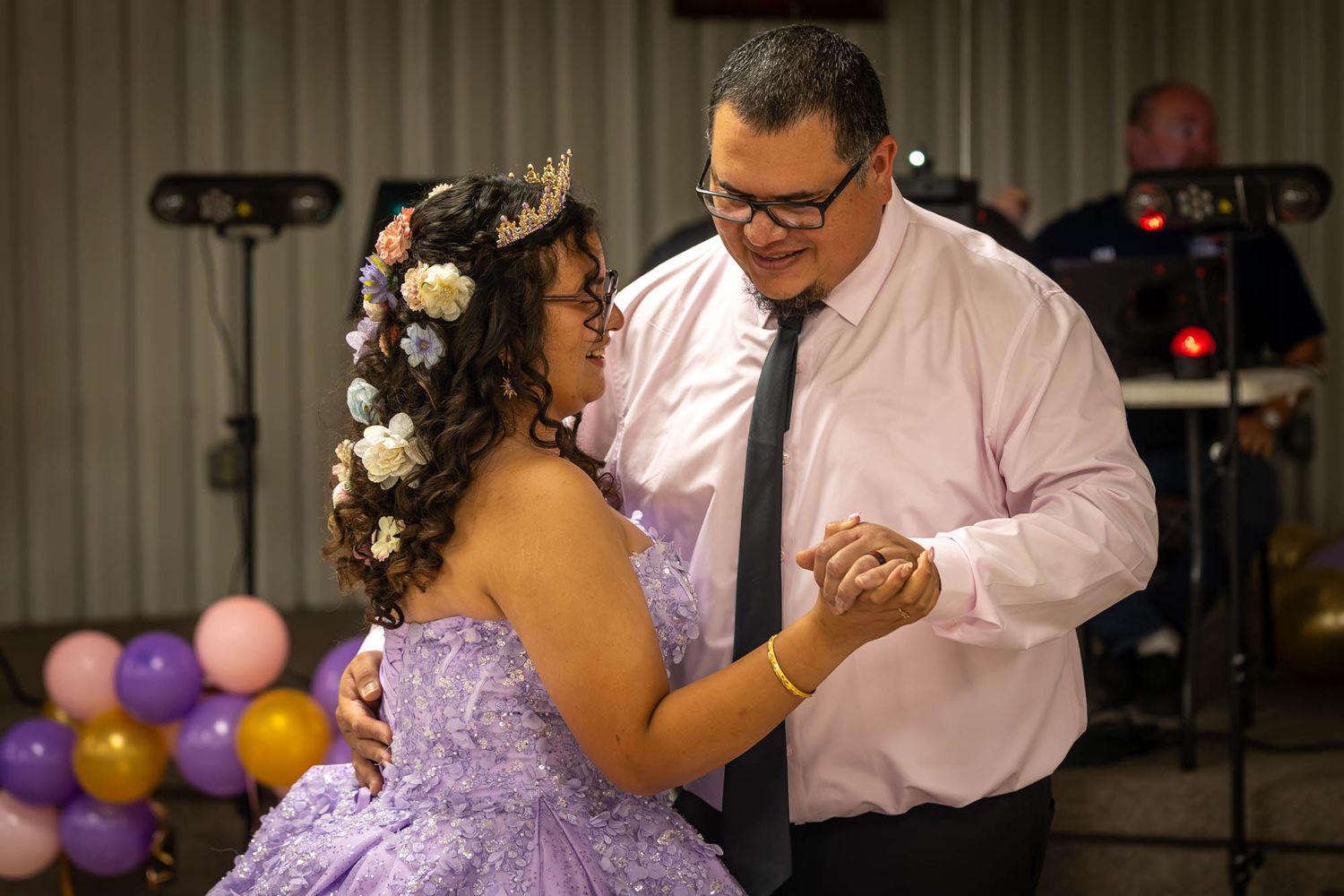 A young woman in a purple quinceañera gown dances with a man in a pink shirt and tie at her celebration.