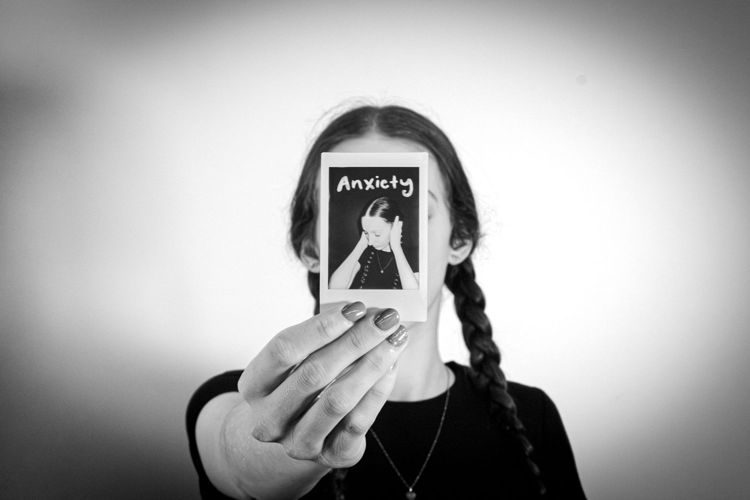 Girl with braids holding a Polaroid labeled Anxiety over her face in a black and white portrait.