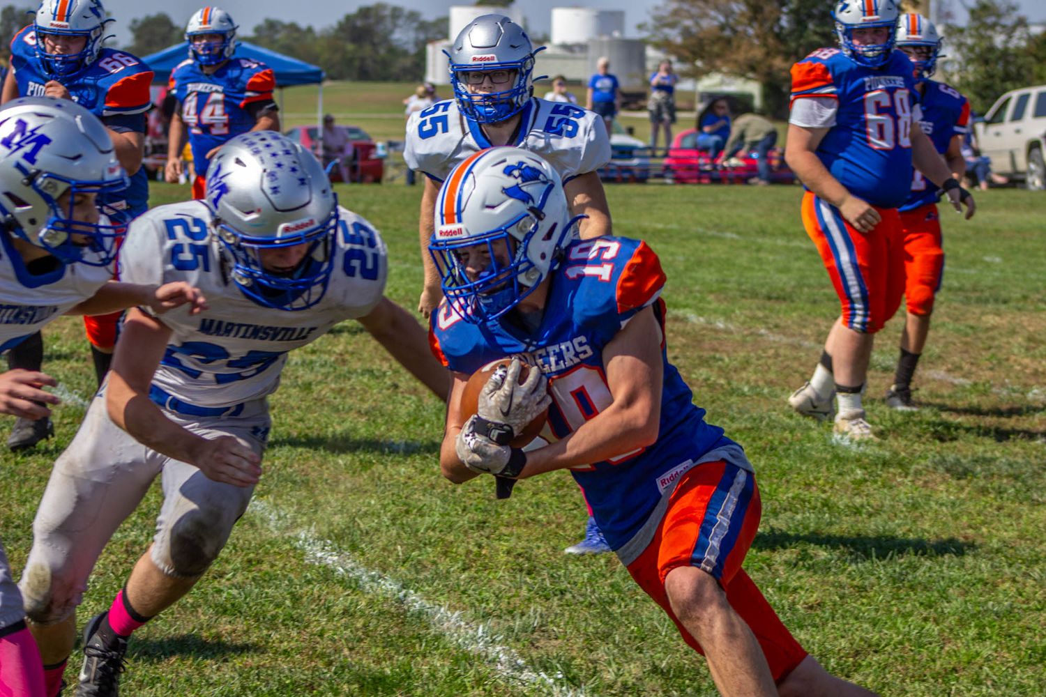 A high school football player in blue and orange uniform runs with the ball while evading opposing defenders.