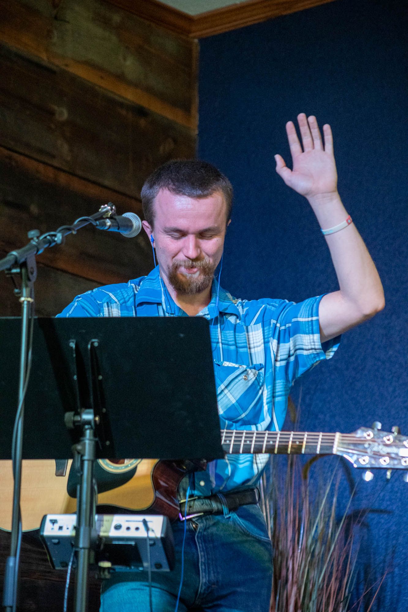 Male musician in blue plaid shirt playing acoustic guitar with one hand raised at an indoor venue.