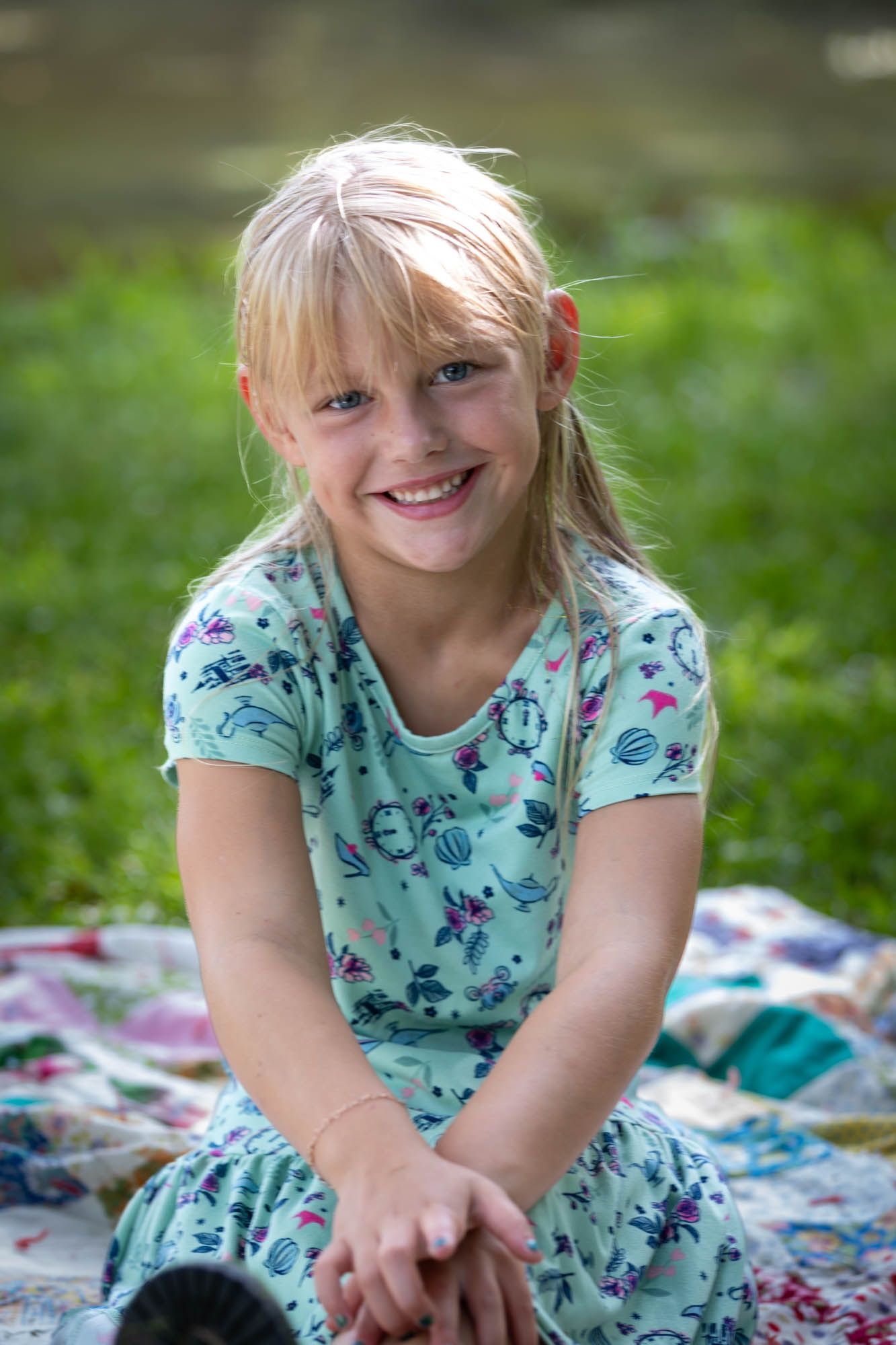 Smiling blonde girl in floral dress sitting on quilt outdoors in green park setting.