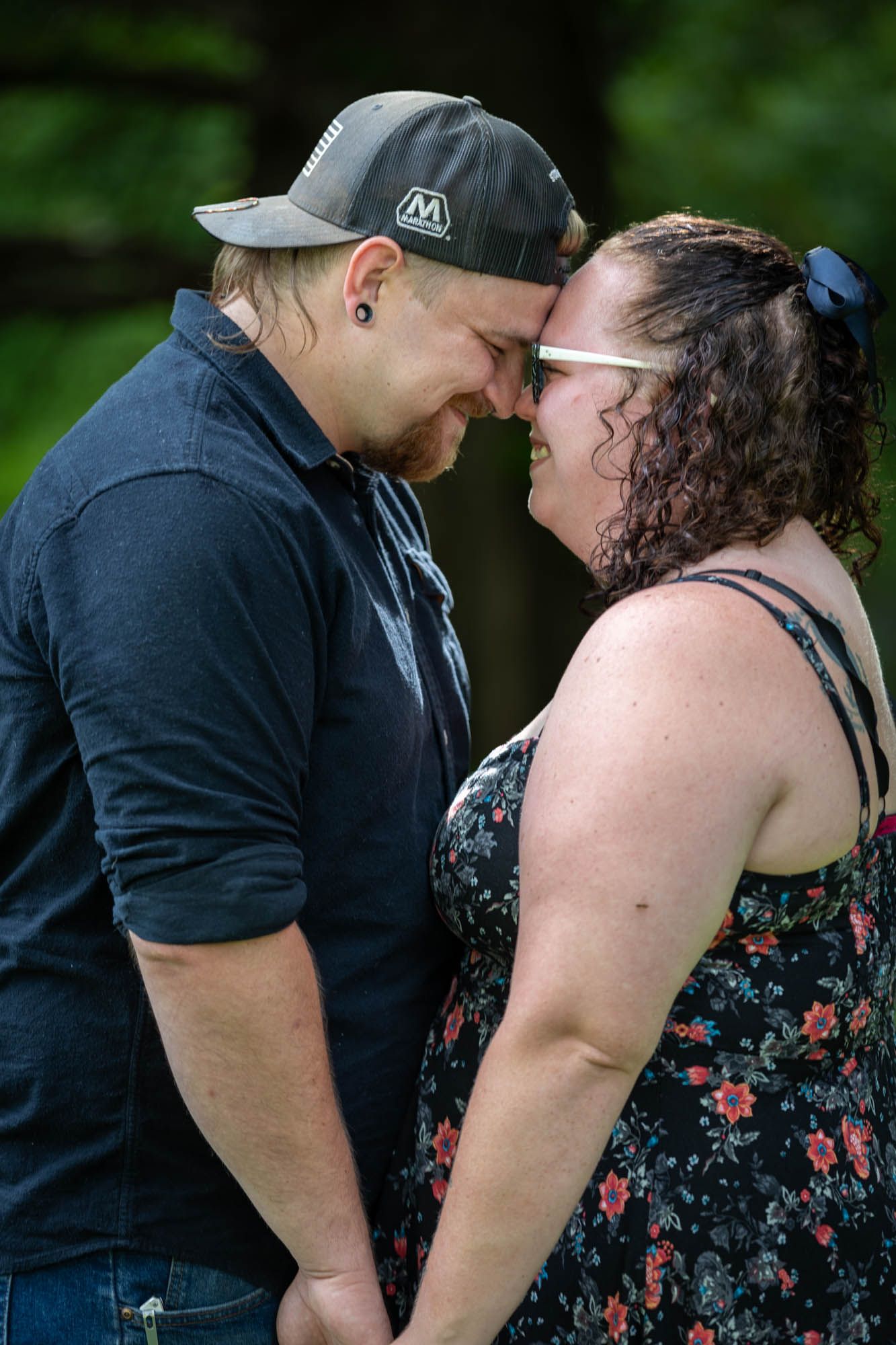 Couple touching foreheads tenderly outdoors, holding hands in a romantic pose surrounded by lush green trees.