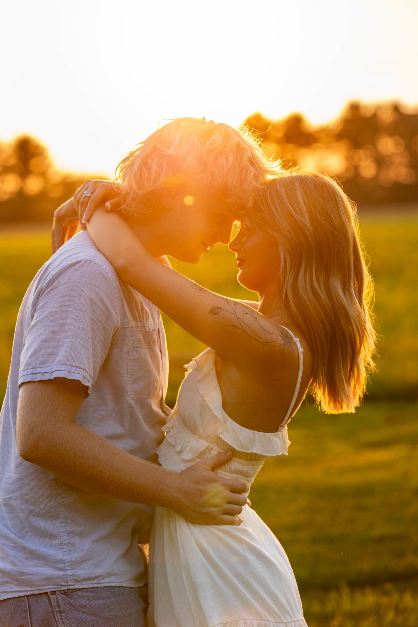 Romantic couple embracing at golden hour sunset in a green field, sharing an intimate moment in warm sunlight.