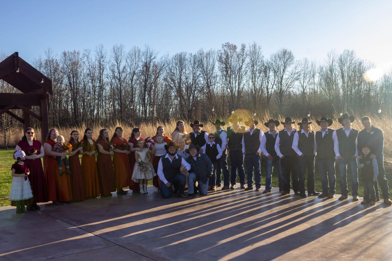 Large wedding party posing outdoors in autumn, bridesmaids in colorful gowns and groomsmen in black suits.