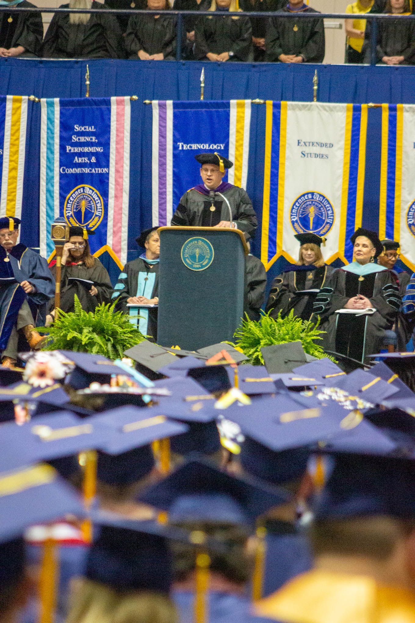 University president speaks at podium during commencement ceremony before graduating students in blue caps.