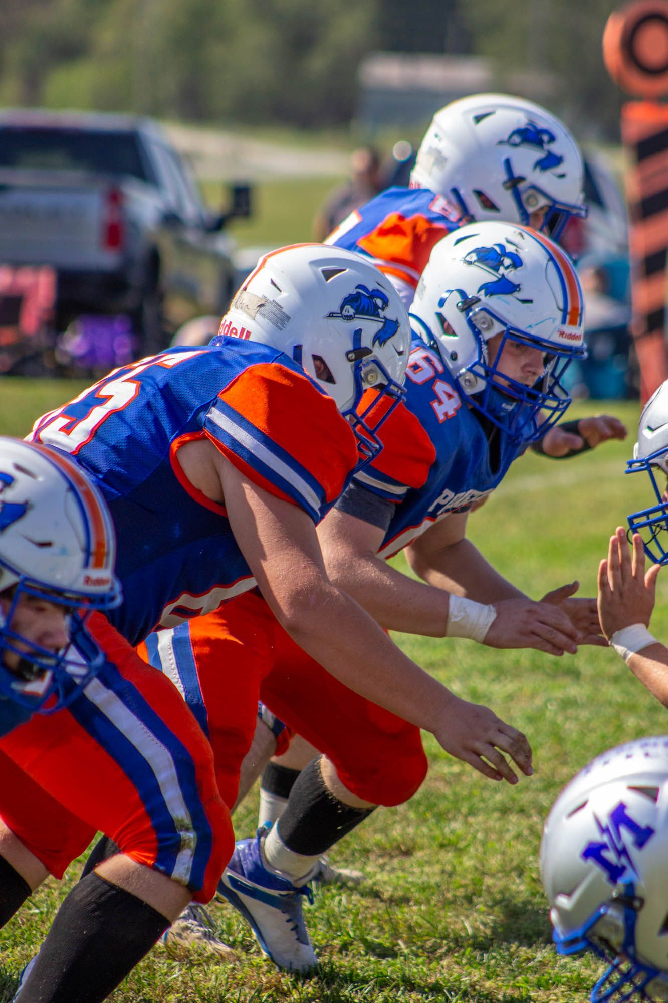 High school football players in blue and orange uniforms engage at the line of scrimmage during a daytime game.