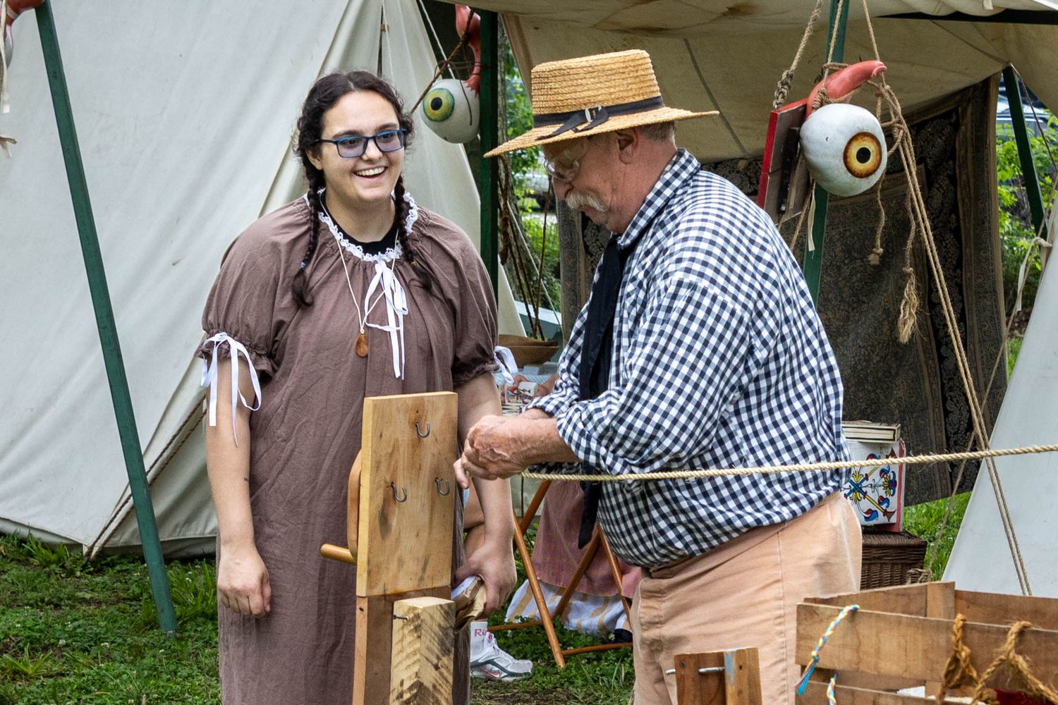 A woman in colonial dress smiles while a man in a straw hat demonstrates rope-making at a historical fair.