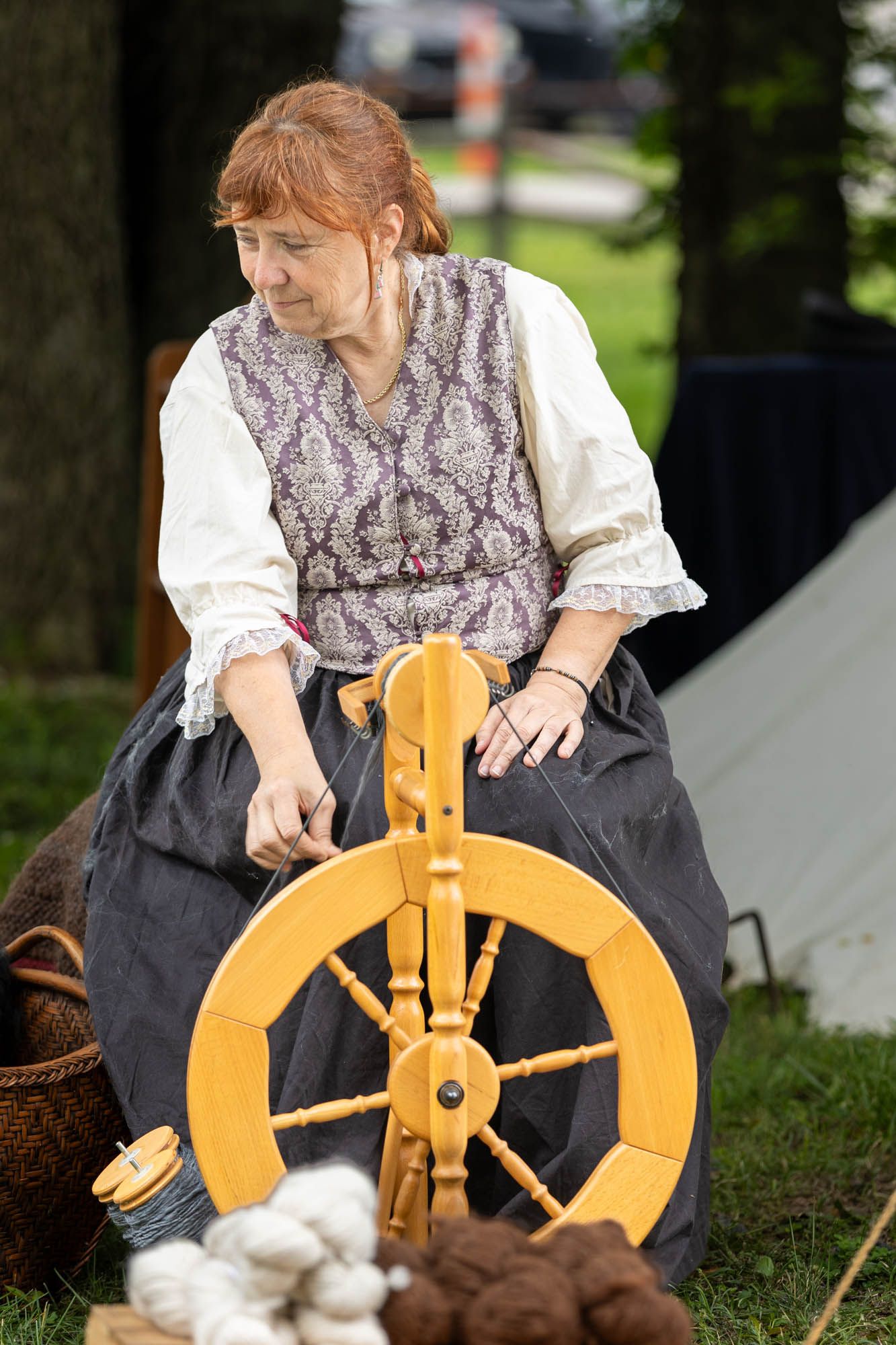 A woman in colonial period costume operates a wooden spinning wheel at an outdoor historical reenactment event.