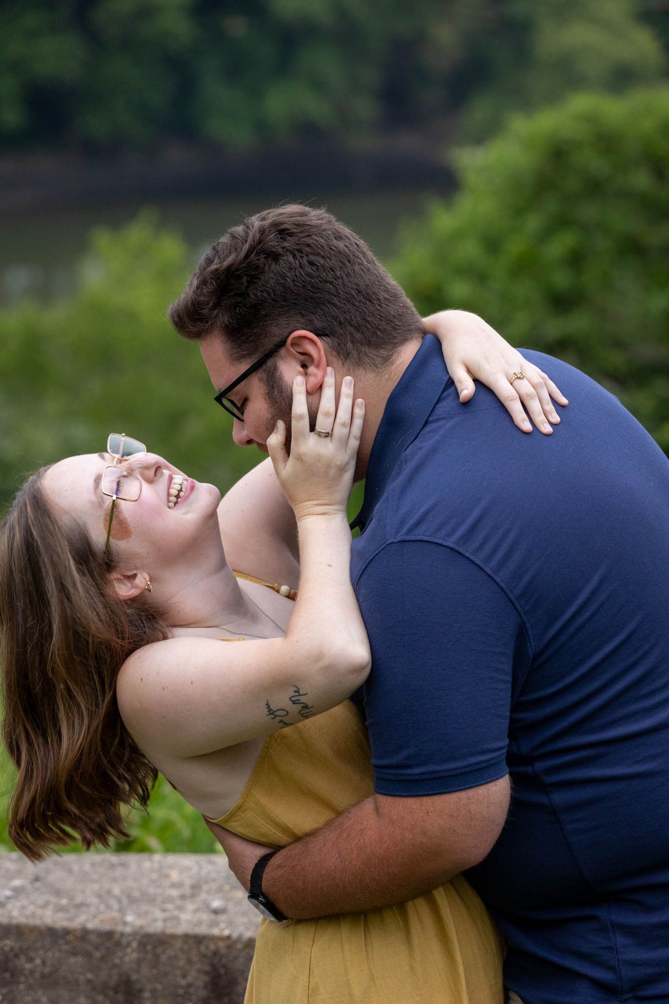 A laughing couple sharing a romantic embrace outdoors, woman in yellow dress with tattoo, man in navy shirt wearing glasses.
