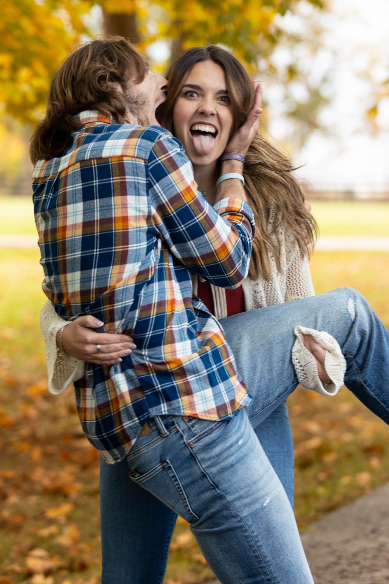 Man lifts laughing woman in his arms during playful outdoor fall engagement photo session.