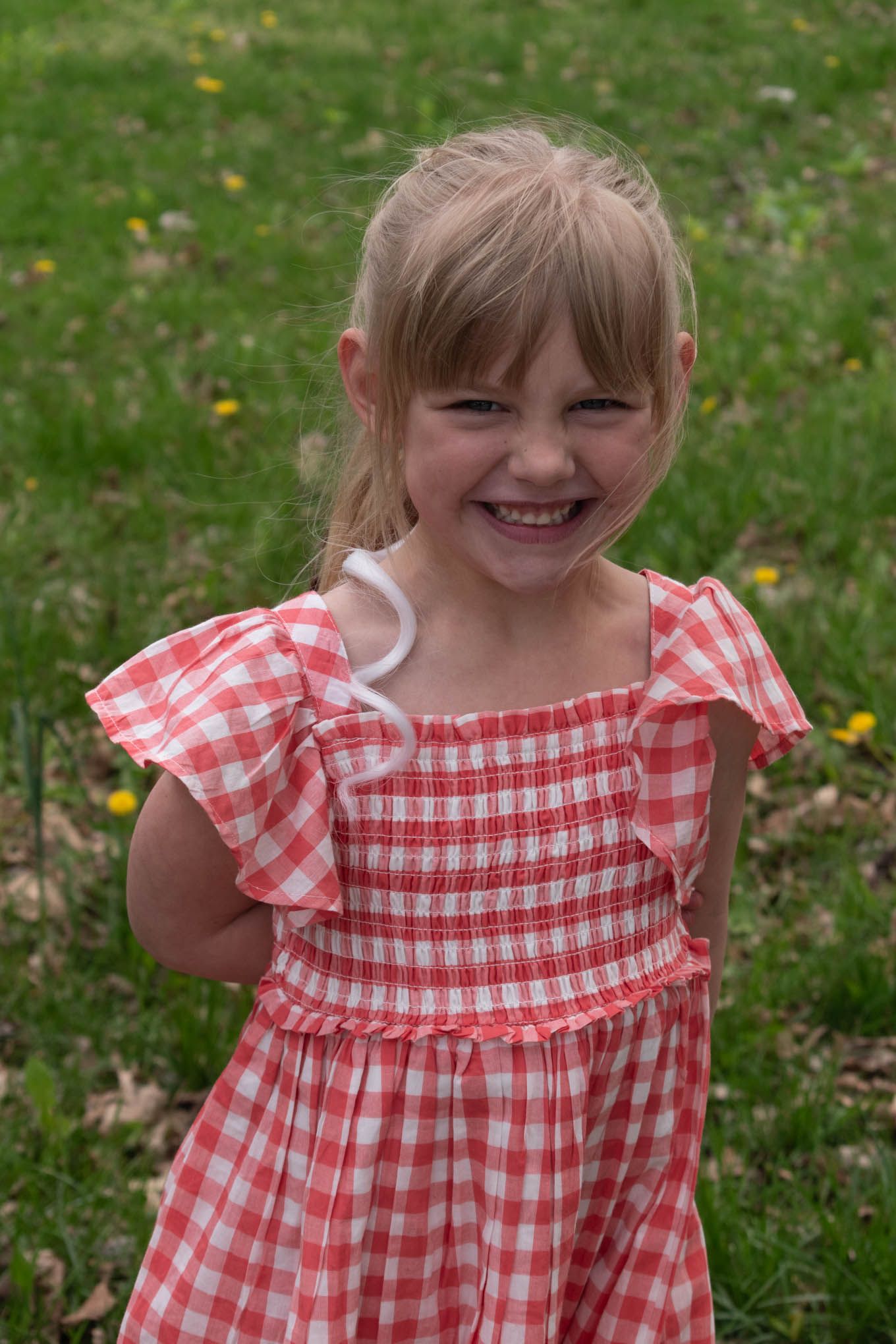 Happy young girl in a red gingham dress laughs joyfully while standing on a green lawn.