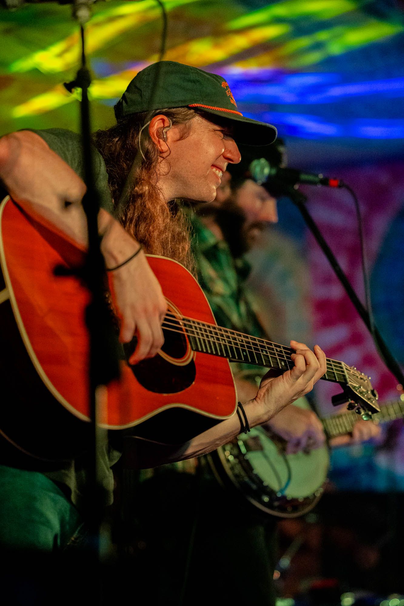 Smiling musician with long hair plays acoustic guitar on a colorful tie-dye decorated stage.