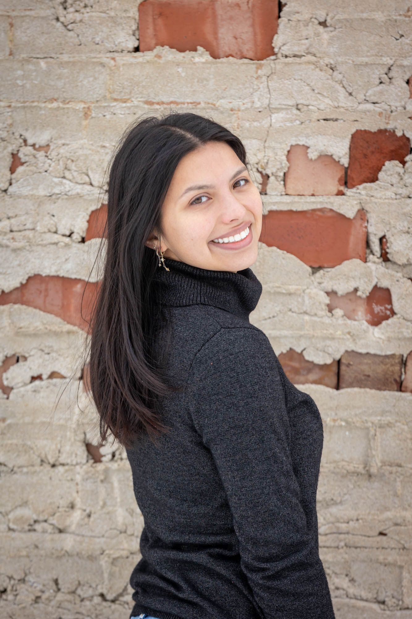 Young woman with long dark hair smiling over her shoulder while leaning against a rustic brick wall.