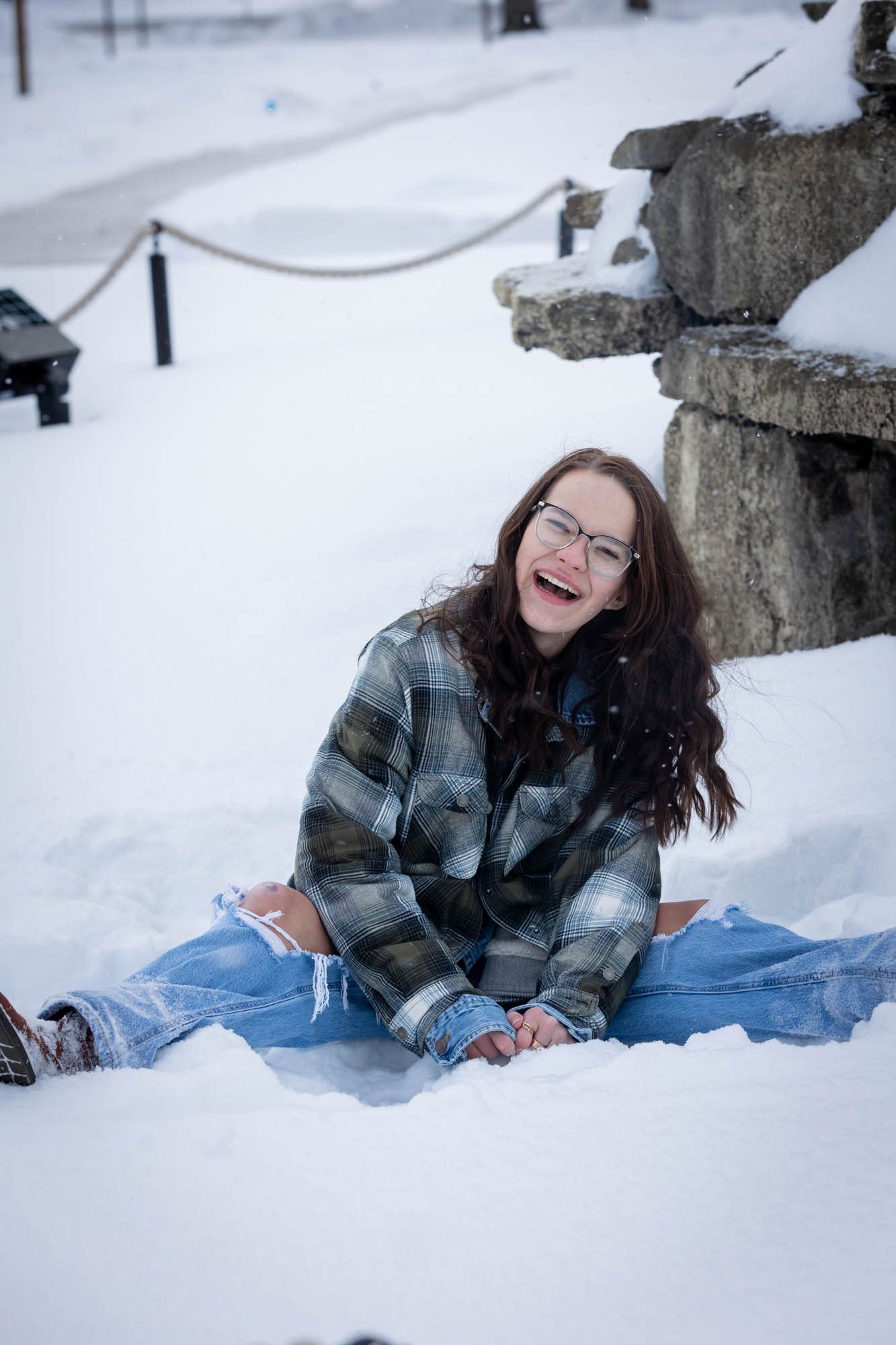 Laughing young woman with glasses sitting in the snow wearing a plaid shirt and ripped jeans outdoors.