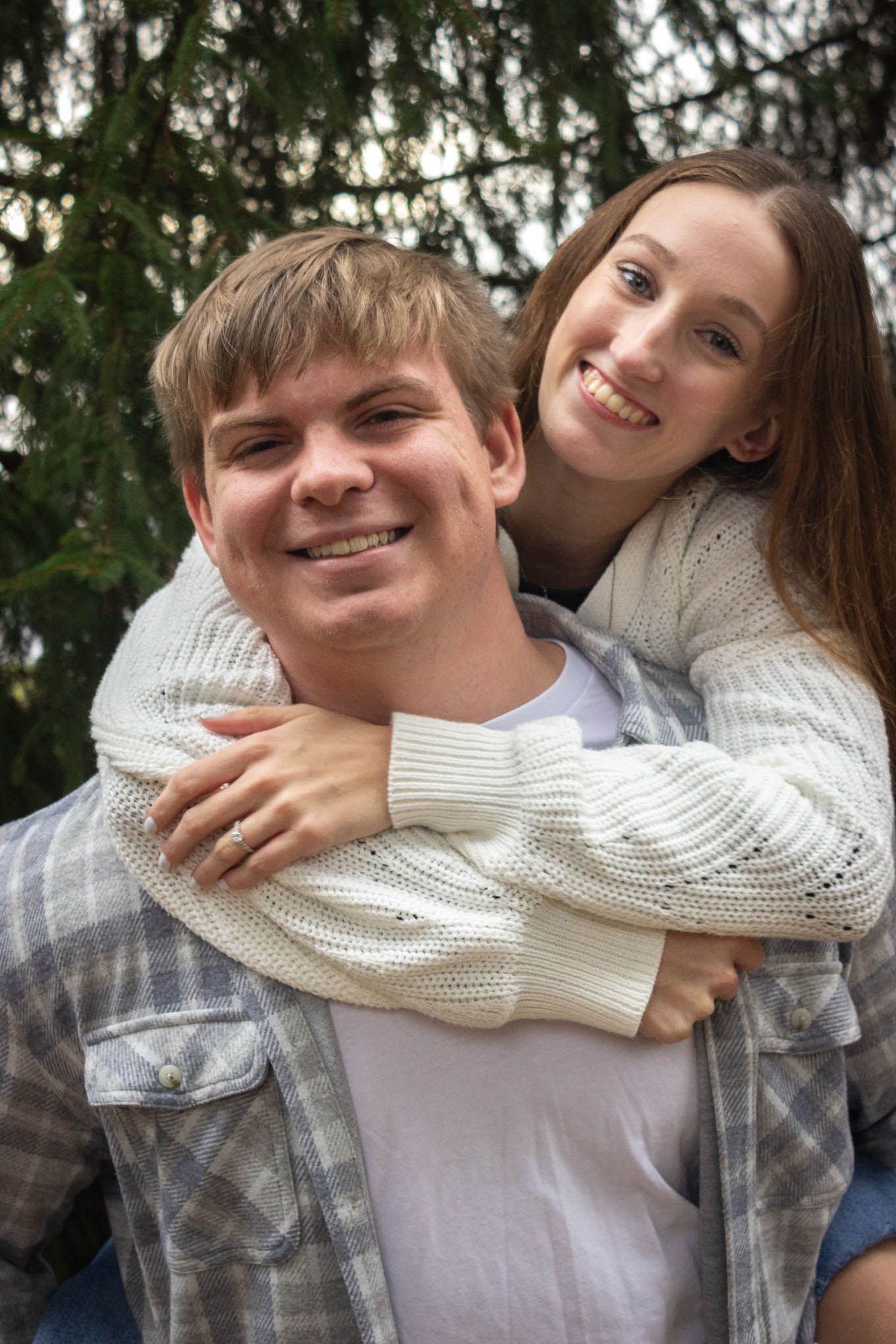 Happy young couple smiling outdoors, woman hugging man from behind near evergreen trees.