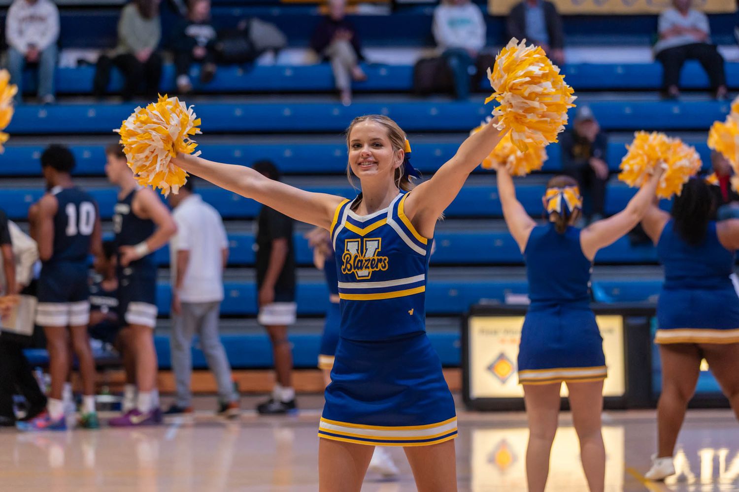 Cheerleader in blue and gold uniform performs with pompoms at a college basketball game.