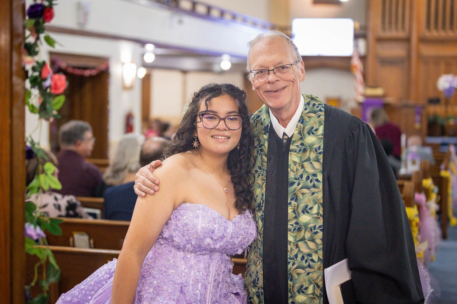 Teenage girl in lavender dress poses with older man in clergy stole inside decorated church.