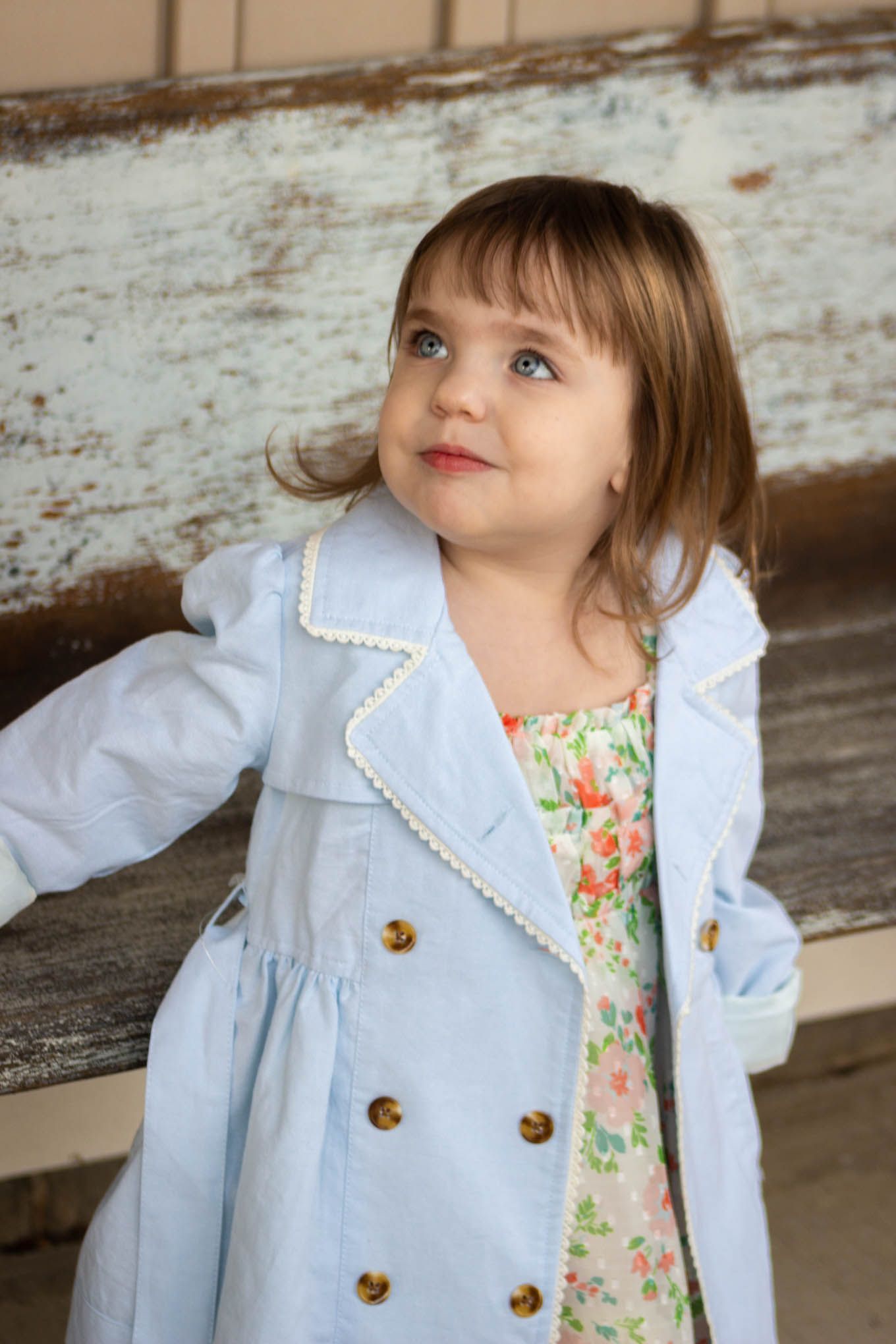 Sweet toddler girl in a light blue double-breasted coat posing against a rustic weathered wooden bench.