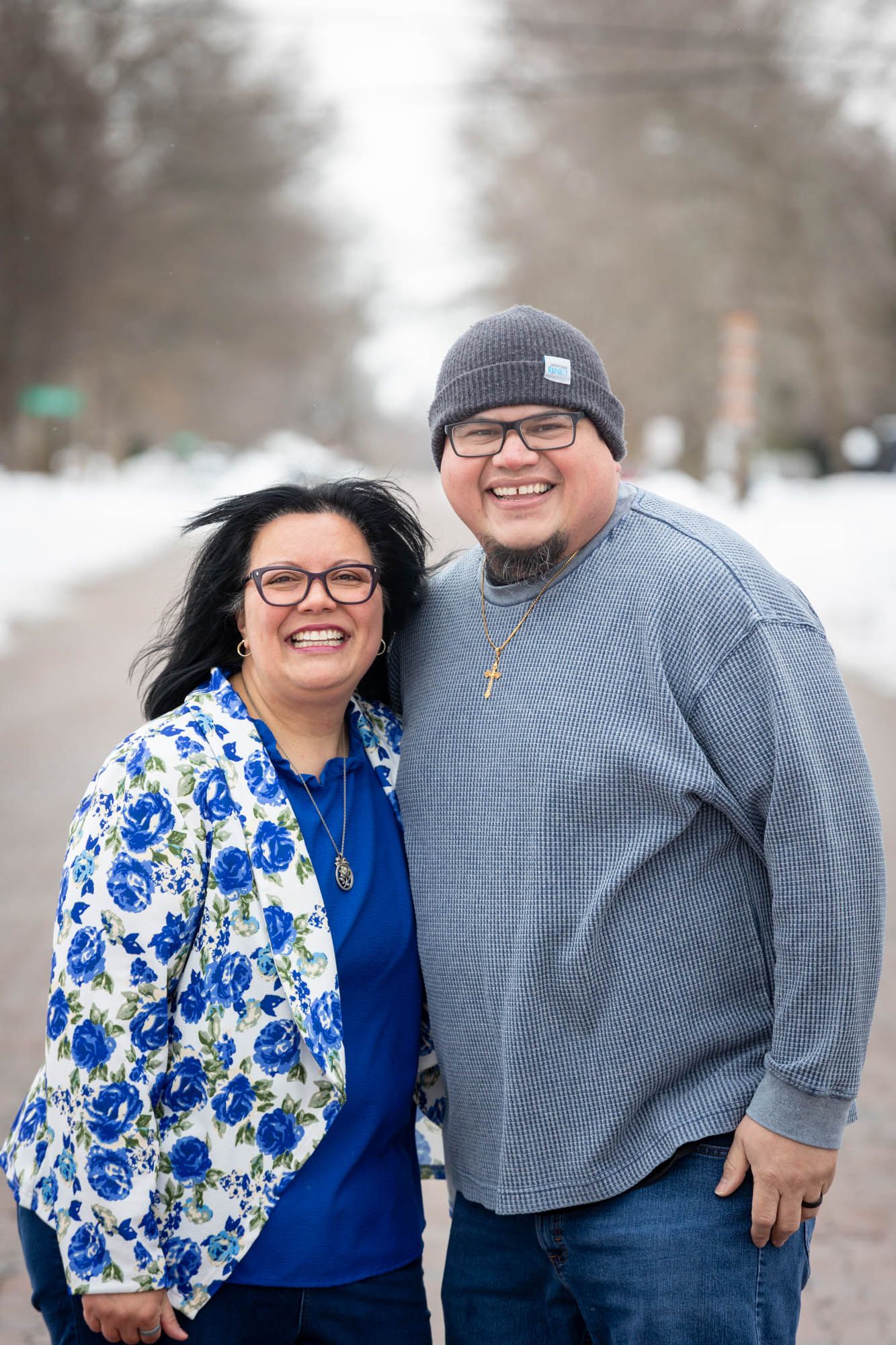 Smiling couple posing together outdoors in winter, man wearing a cross necklace and gray beanie hat.
