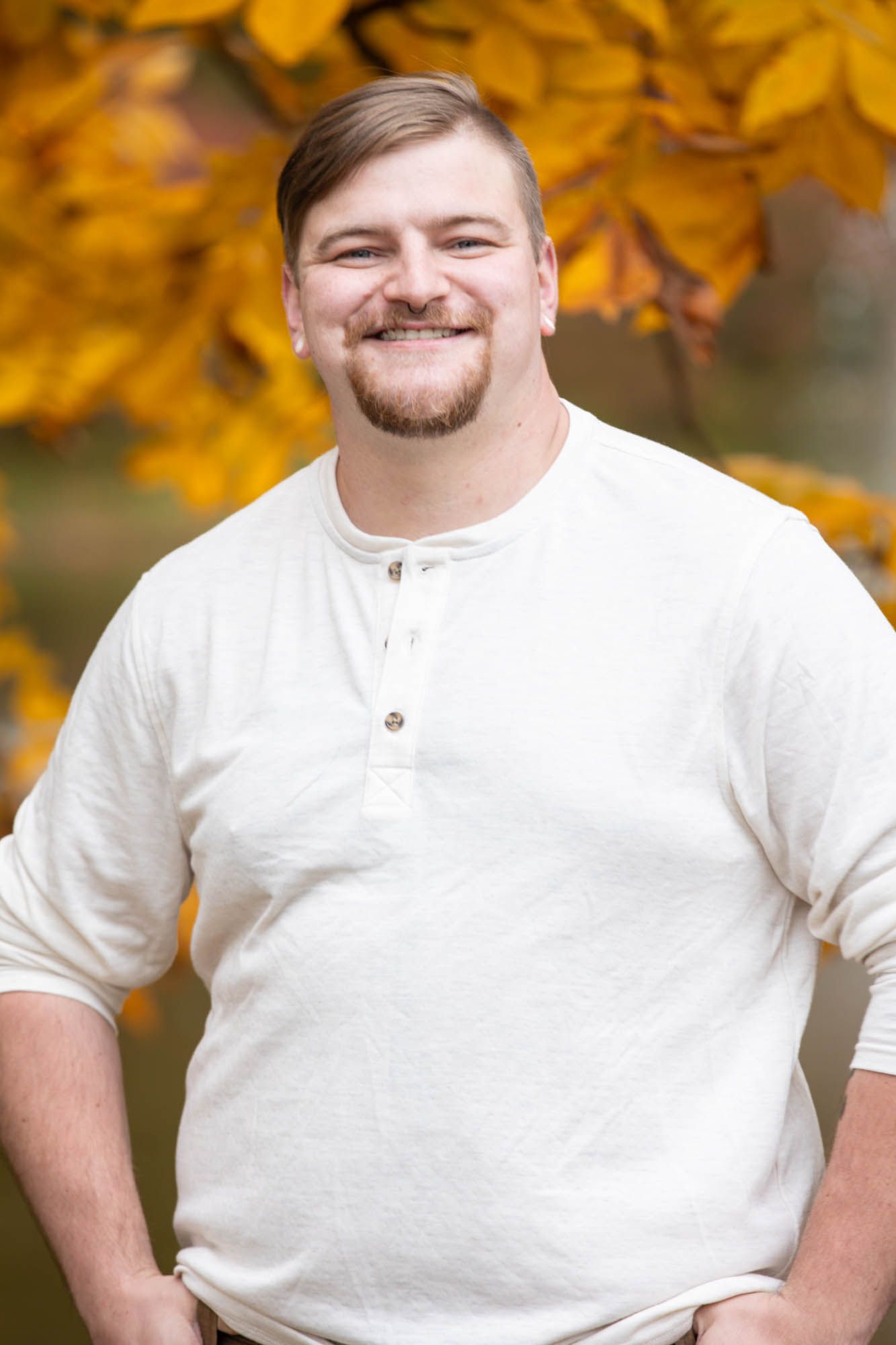 Bearded man smiling warmly in a white henley shirt with golden autumn leaves in the background.