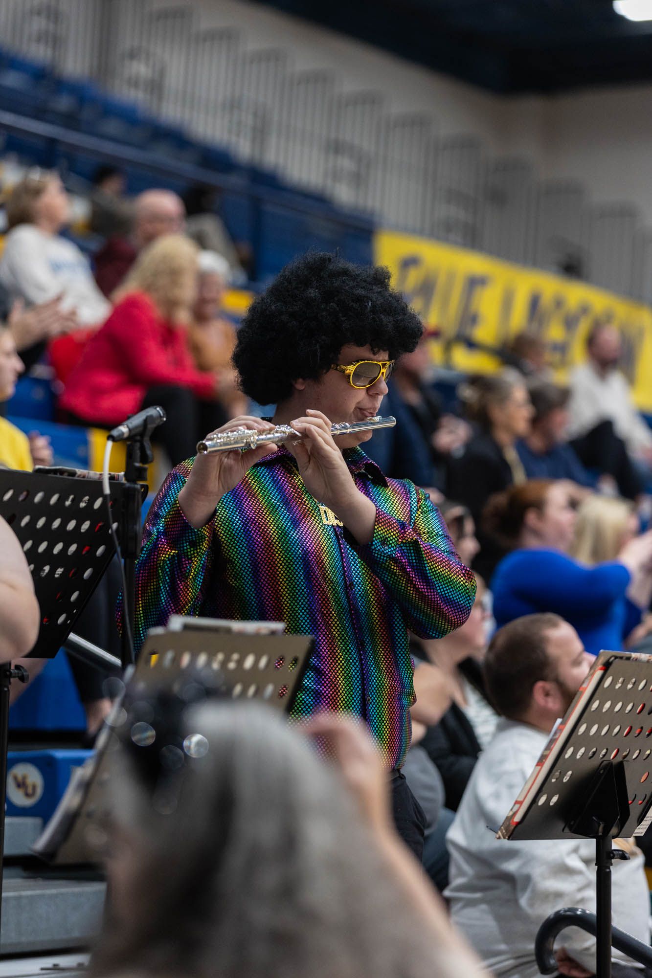 Flutist in colorful disco shirt and afro wig performs in pep band at college basketball game.