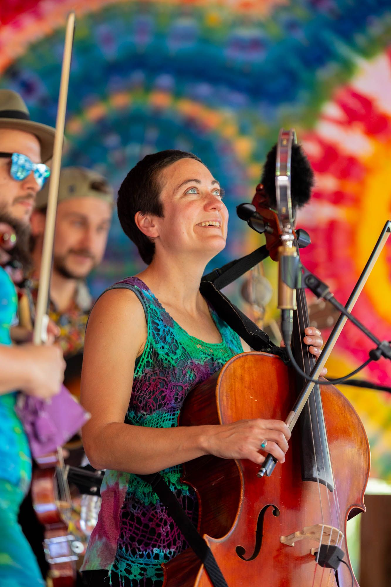 A female cellist smiles while performing on stage at a colorful outdoor music festival.