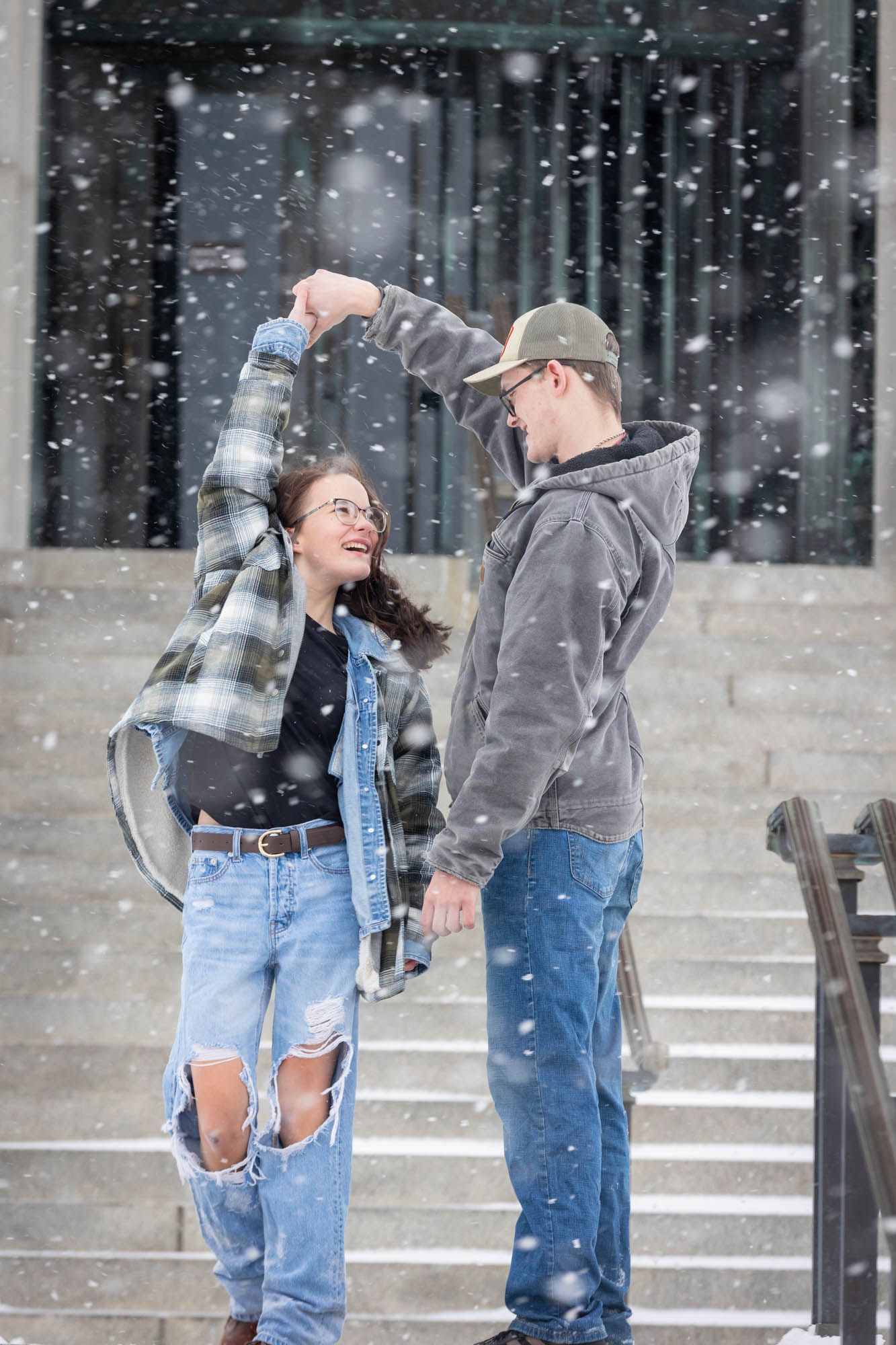 Joyful young couple dancing and spinning together on snowy steps during snowfall.